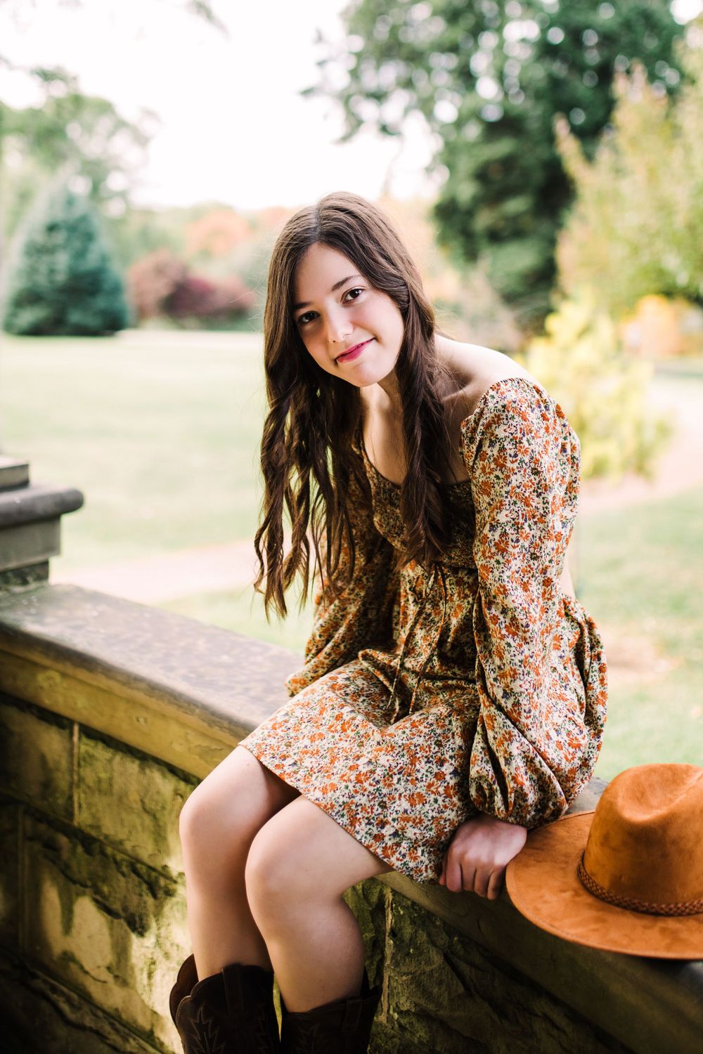 Young woman in floral dress and cowboy boots sitting on stone wall in autumn garden.