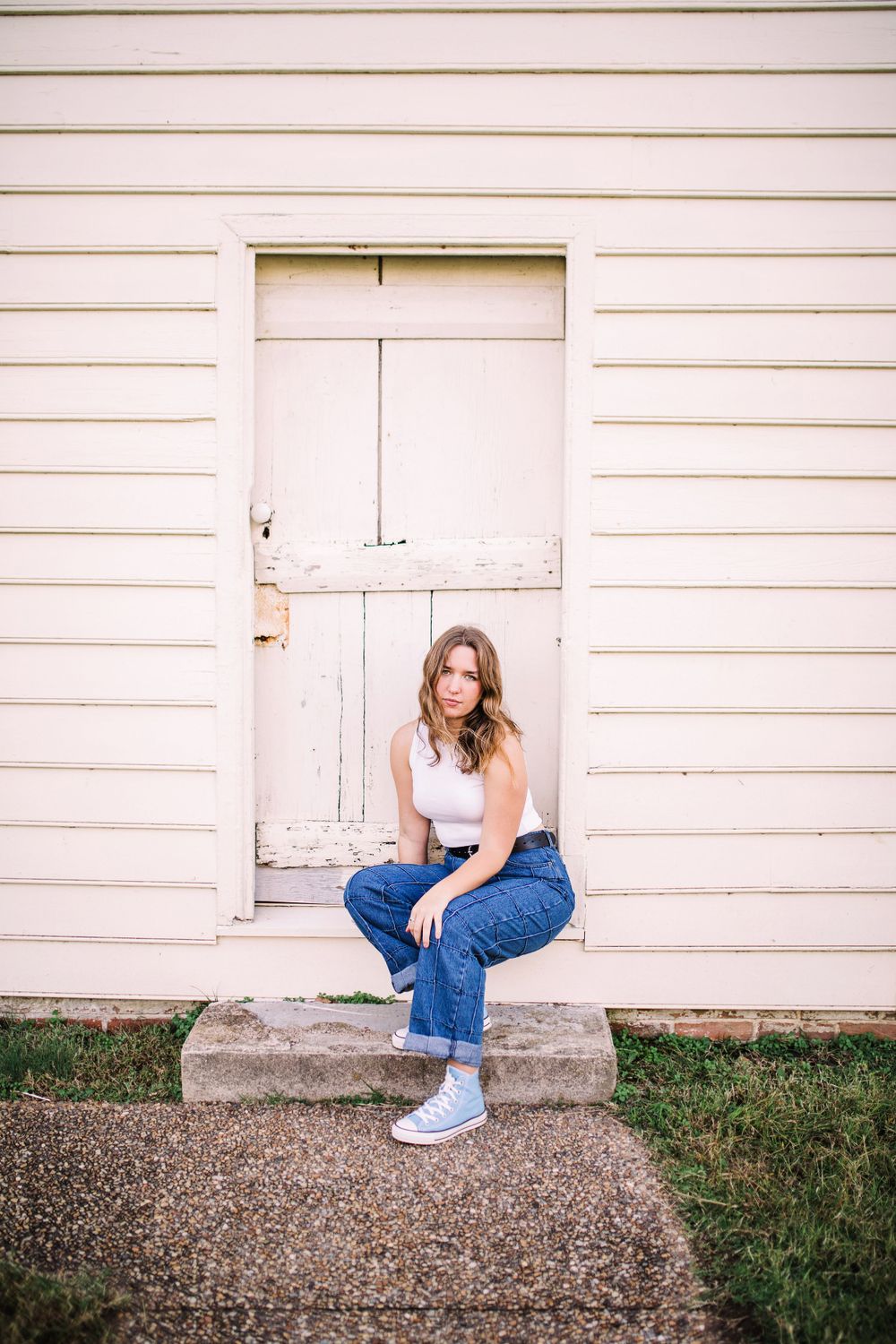 Young woman in white tank top and jeans sitting on steps by a vintage white wooden door.