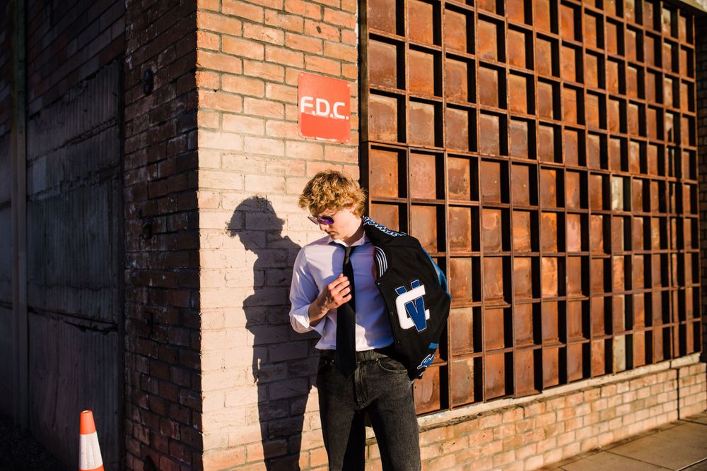 Young man in white shirt and varsity jacket walking past brick wall with dramatic shadow.