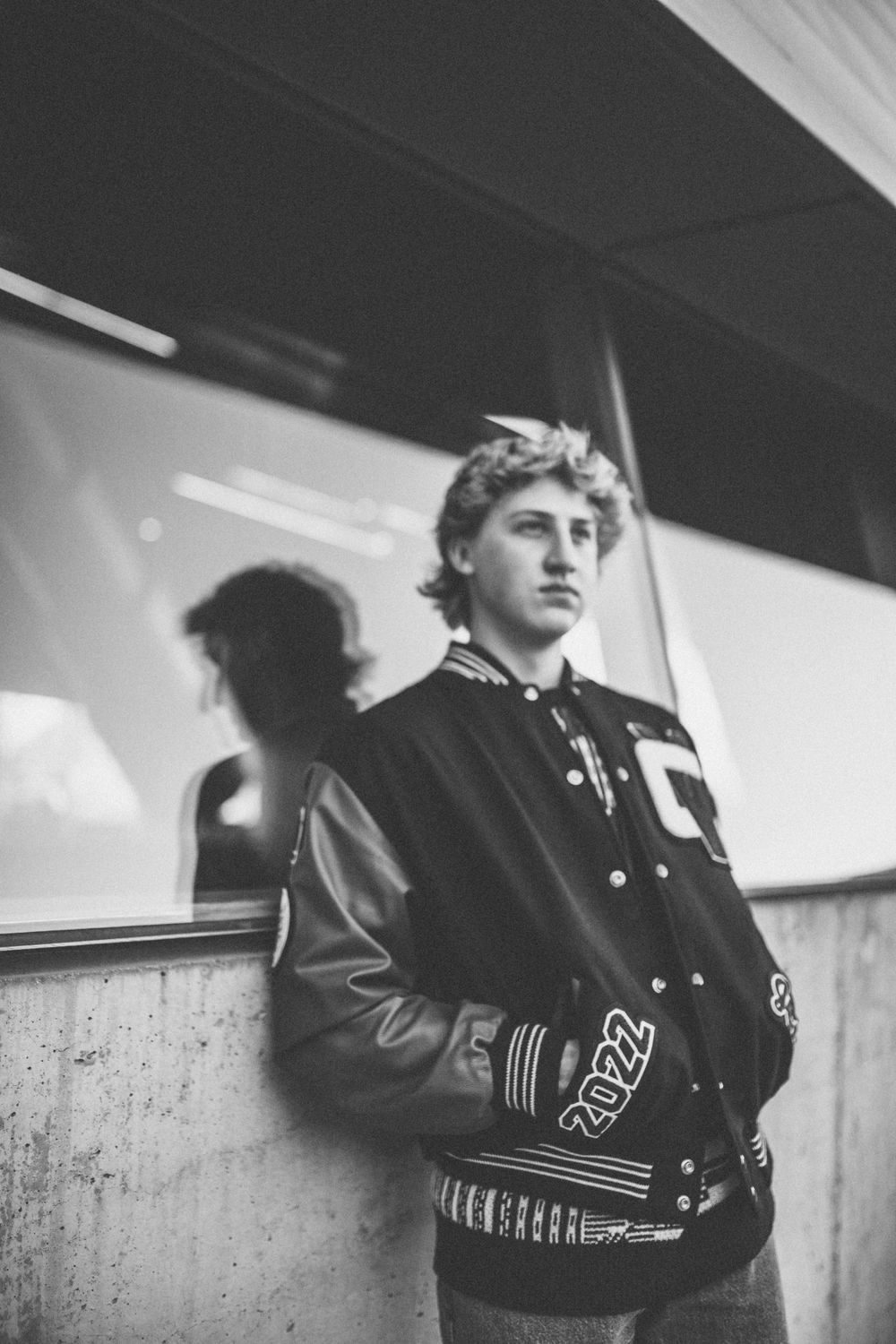 Black and white portrait of teen in 2022 varsity jacket leaning against glass wall.