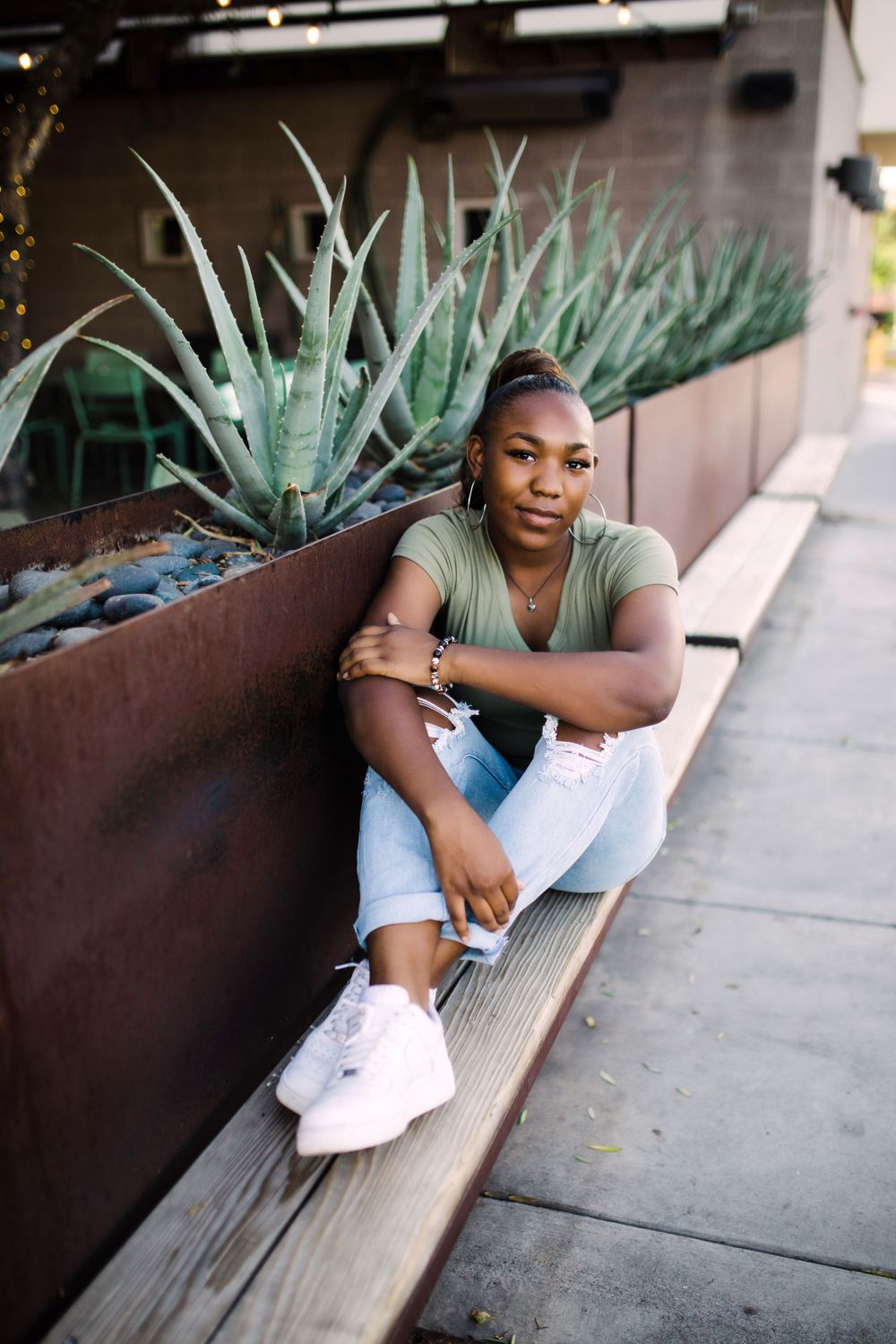 Young Black woman in olive tee and ripped jeans sits against rustic metal planter with agave plants.