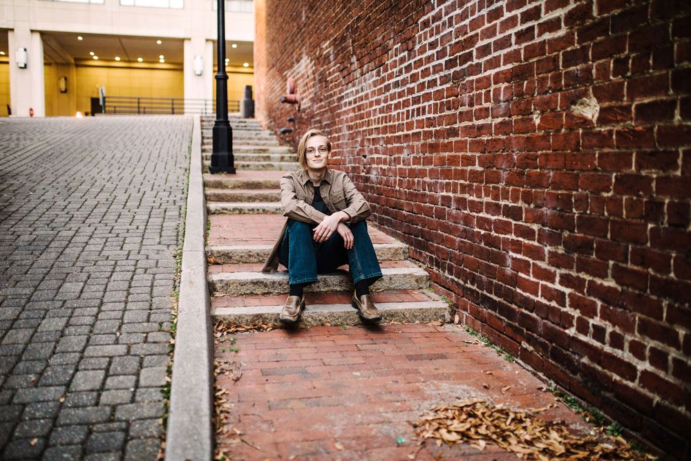 Person in brown jacket sitting on brick steps beside weathered red brick wall in urban alley.