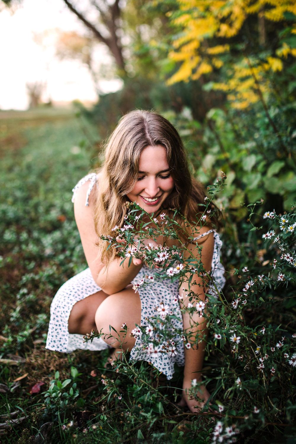 Smiling young woman in white dress crouching among wild pink wildflowers in lush green field.
