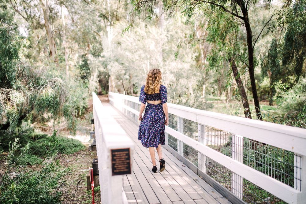 Young woman in navy floral dress walking along a wooden bridge surrounded by trees.