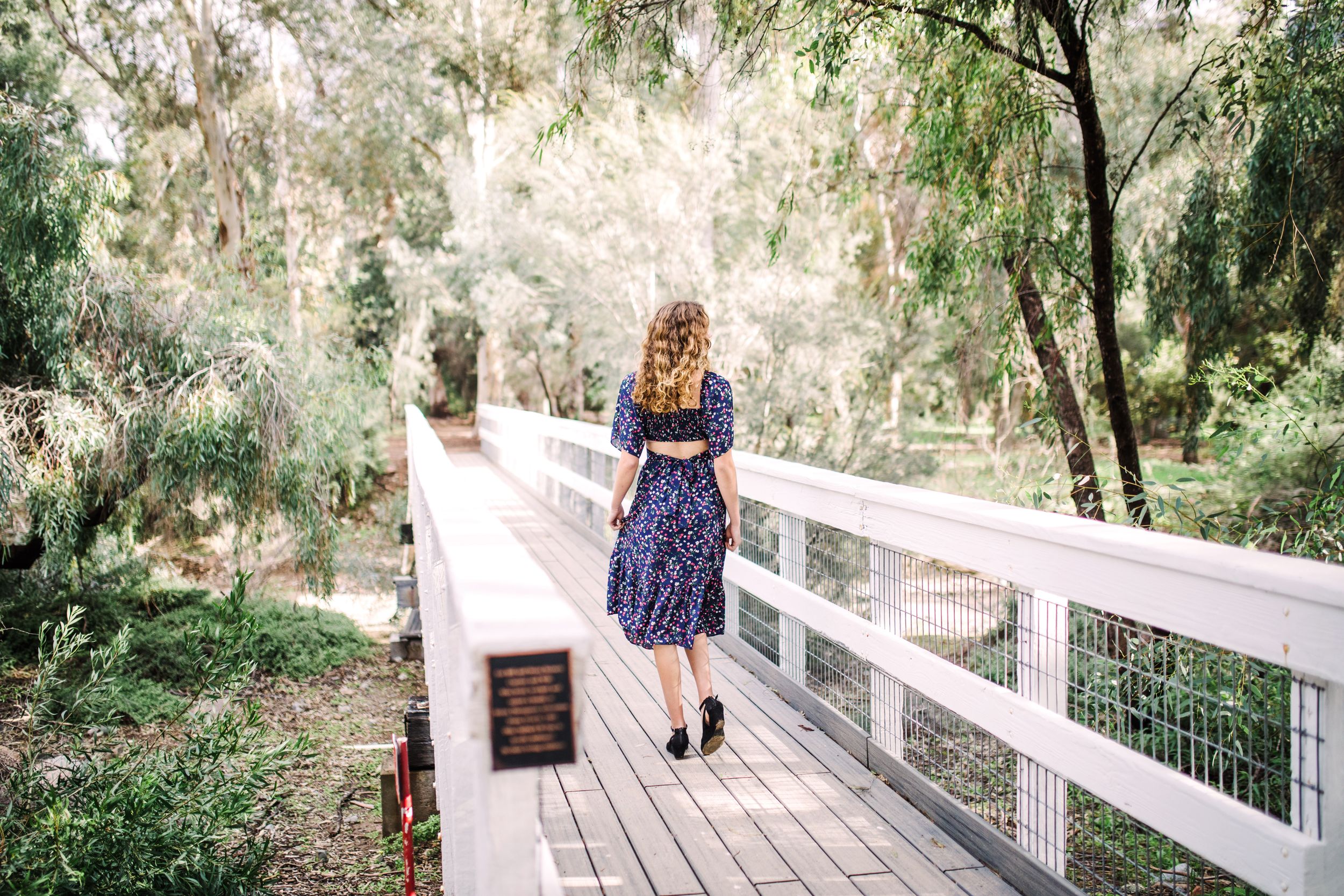 Young woman in navy floral dress walking along a wooden bridge surrounded by trees.