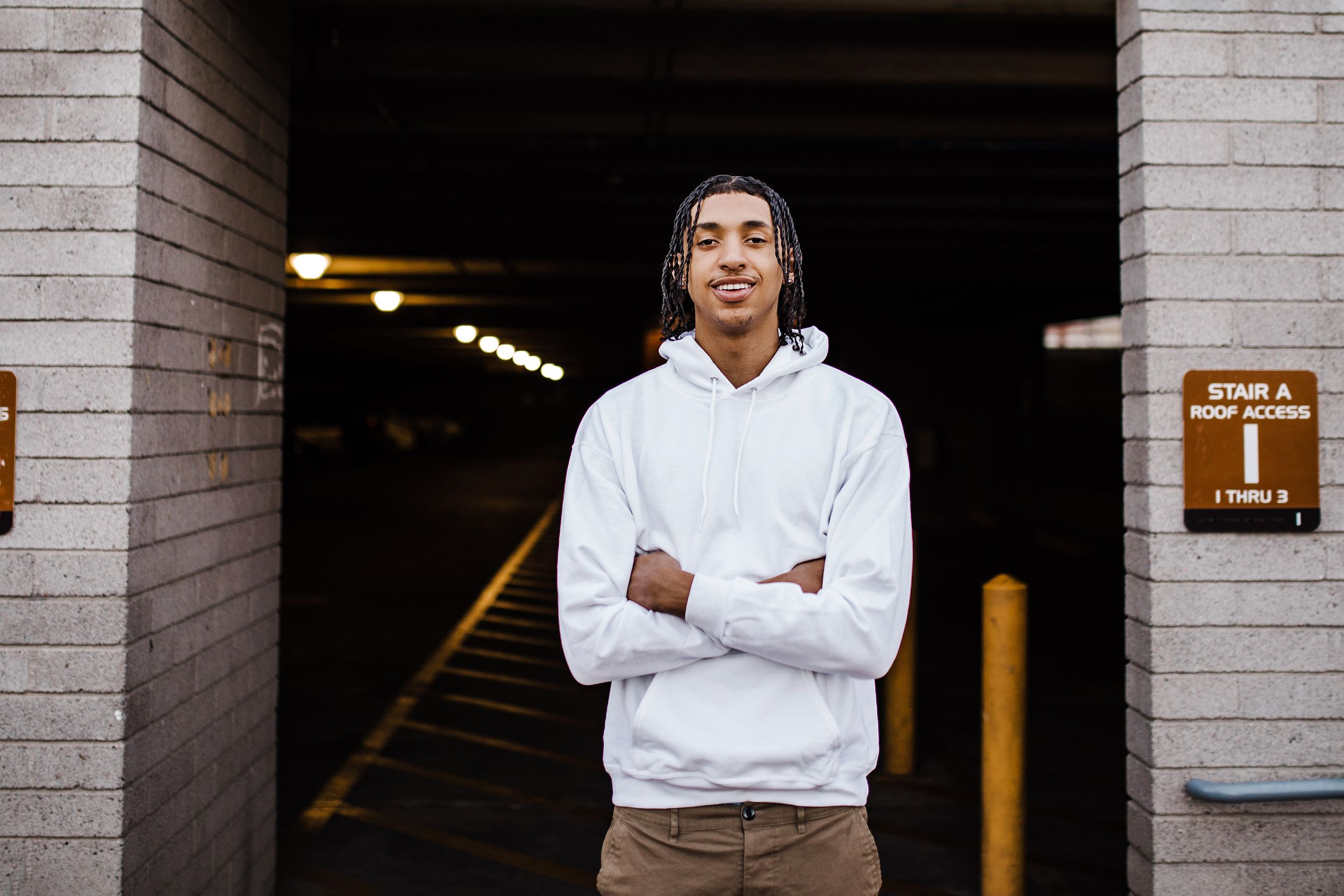 Young man with braids in white hoodie stands with arms crossed at parking garage entrance.