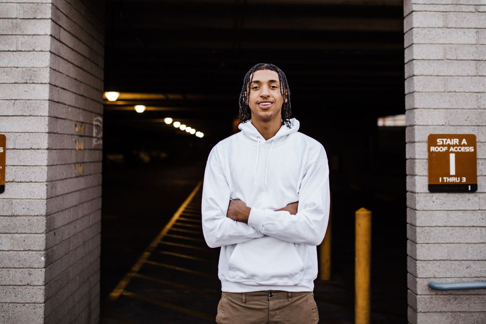 Young man with braids in white hoodie stands with arms crossed at parking garage entrance.