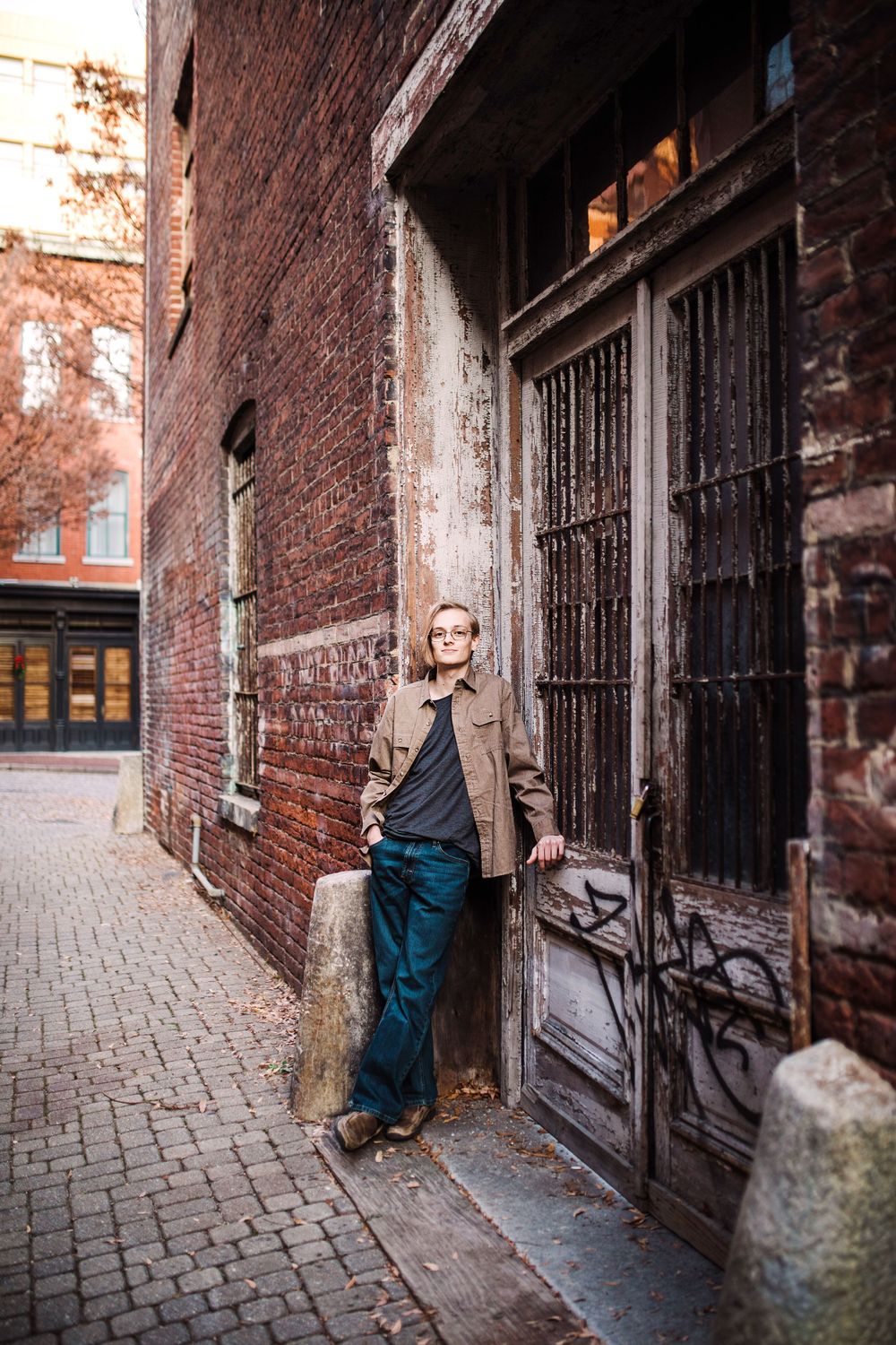 Man in brown jacket leaning against vintage brick building with ornate wooden door in urban alley.