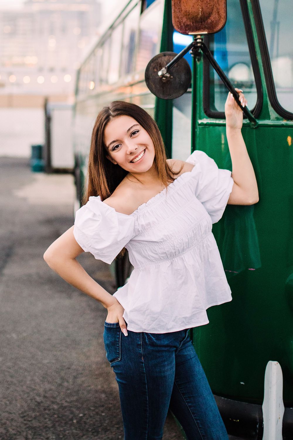 Cheerful young woman in white off-shoulder top posing beside a vintage green bus.