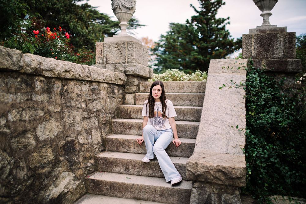 Young woman in white tee and flare jeans sitting on stone garden steps surrounded by greenery.
