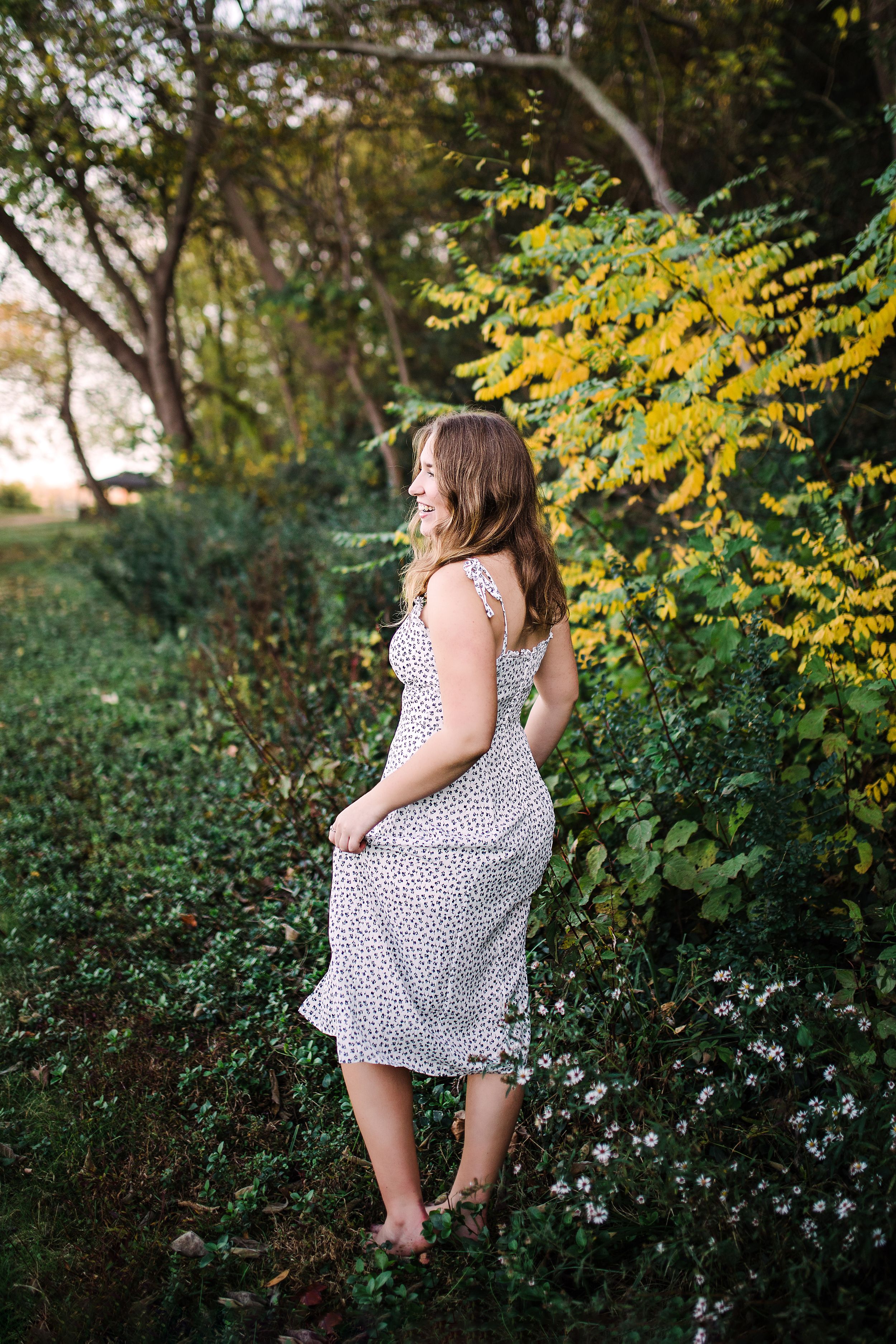 Young woman in white floral midi dress standing among green and yellow foliage in a natural outdoor setting.