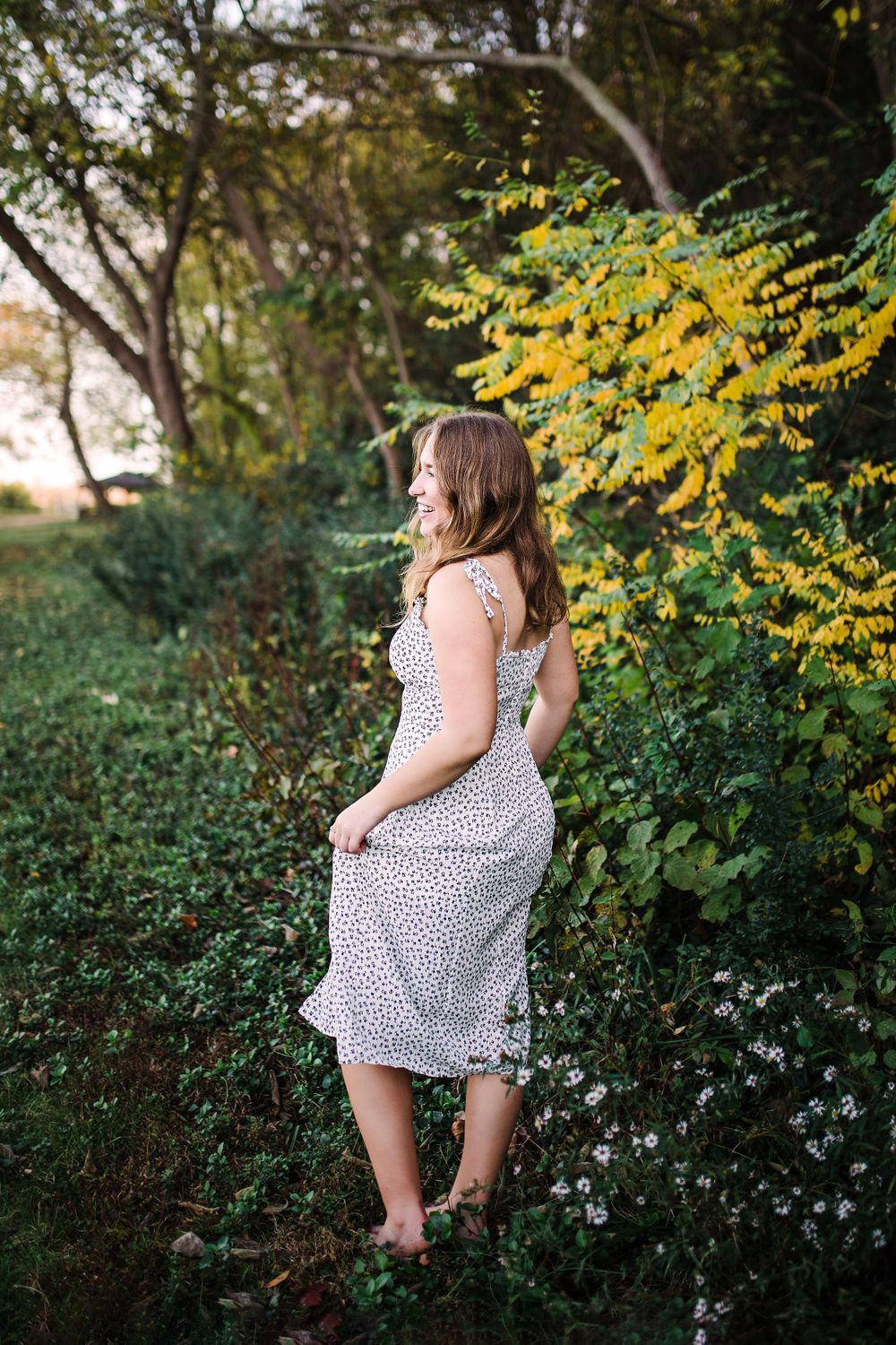 Young woman in white floral midi dress standing among green and yellow foliage in a natural outdoor setting.
