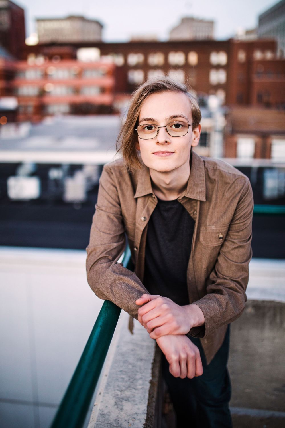 Young person with glasses leaning on rooftop railing wearing brown jacket with urban cityscape background.