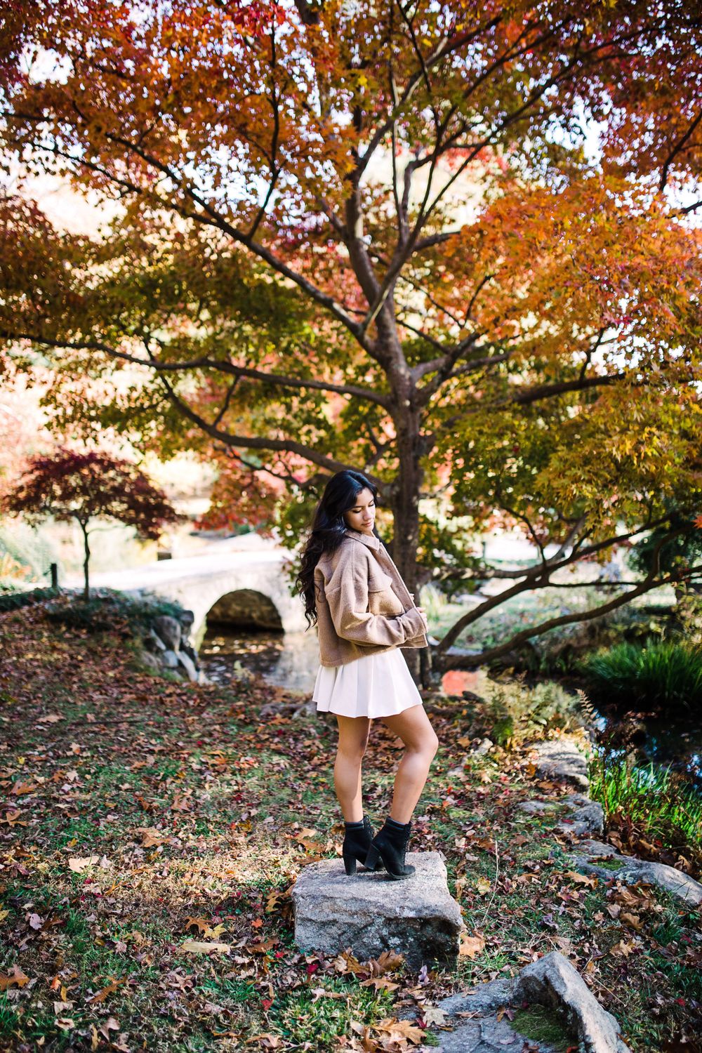 Young woman in tan jacket and white skirt stands on rocks beneath vibrant autumn foliage.