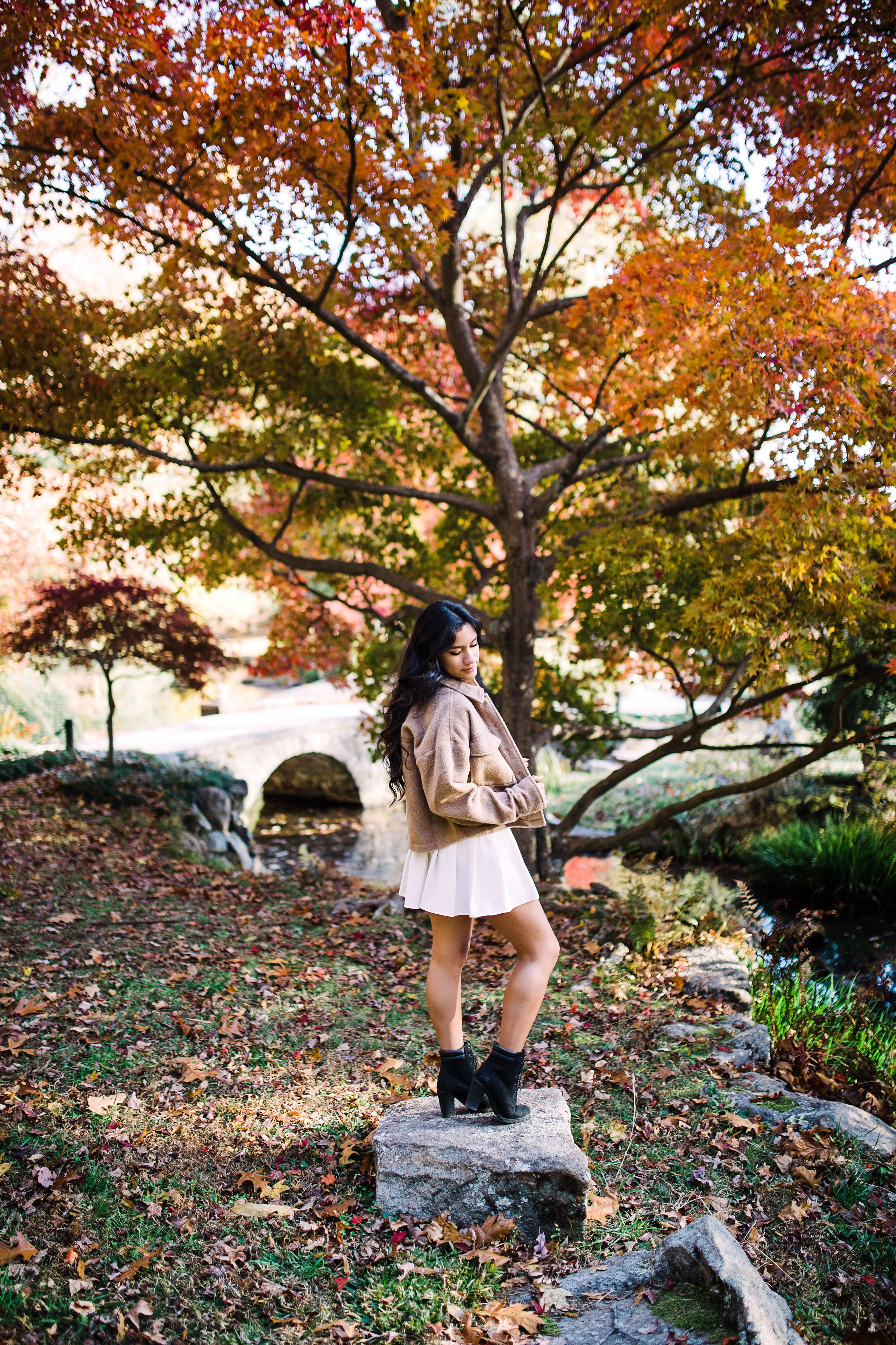 Young woman in tan jacket and white skirt stands on rocks beneath vibrant autumn foliage.