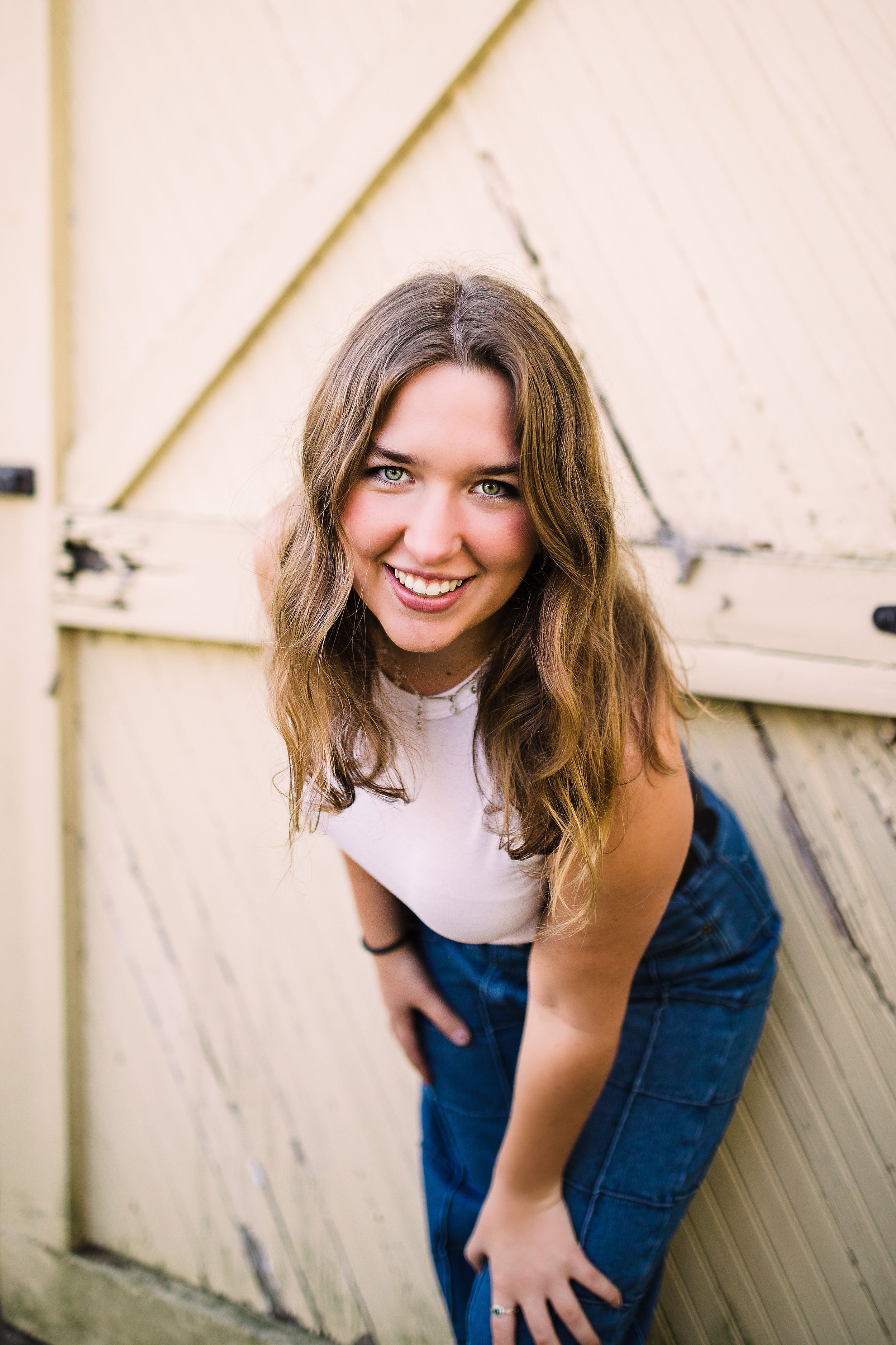 Smiling young woman in white crop top and denim leaning forward against a rustic pale yellow barn door.