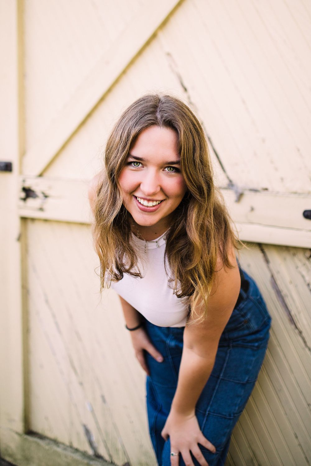 Smiling young woman in white crop top and denim leaning forward against a rustic pale yellow barn door.