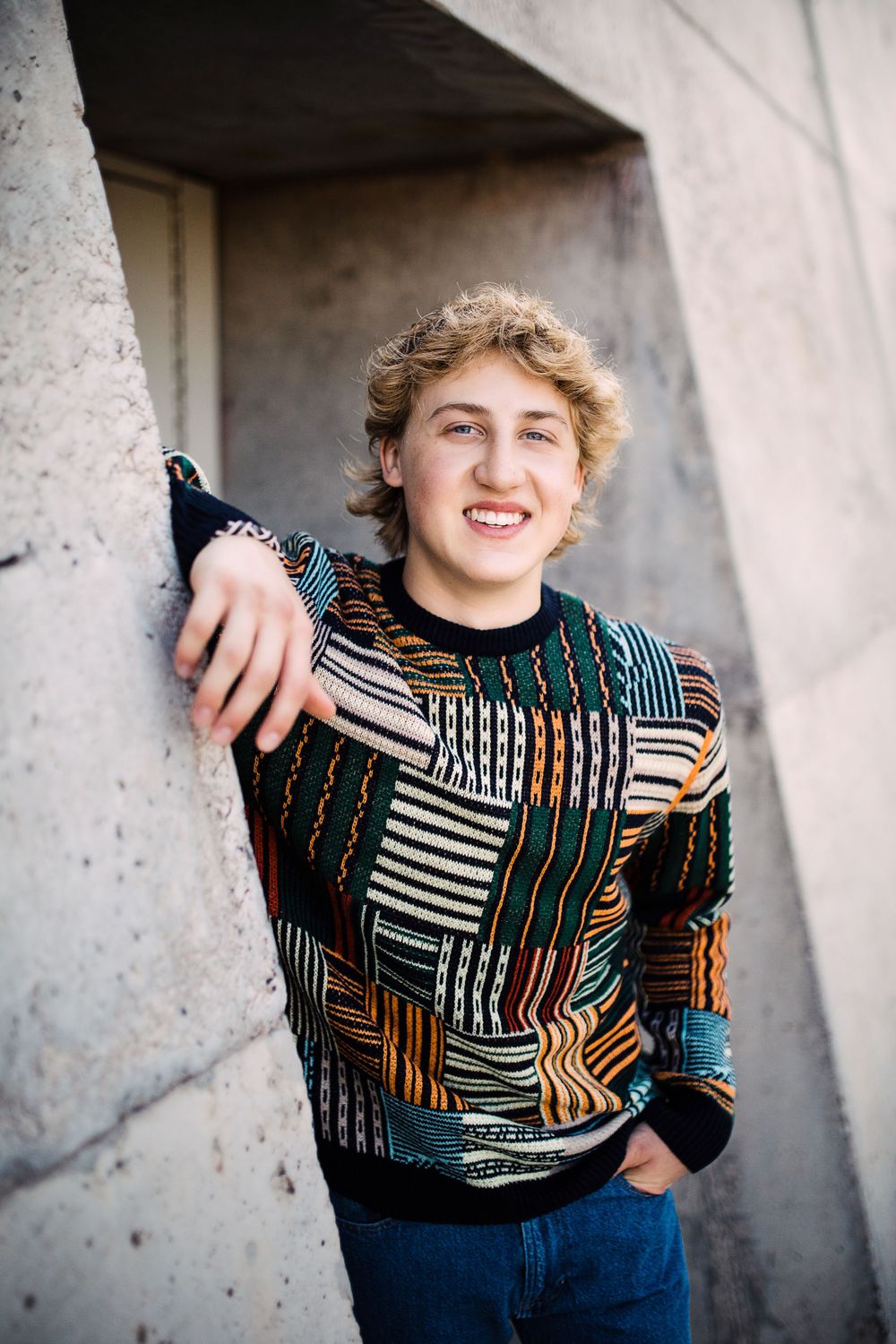 Smiling teen boy in colorful patchwork sweater leaning against concrete wall outdoors.