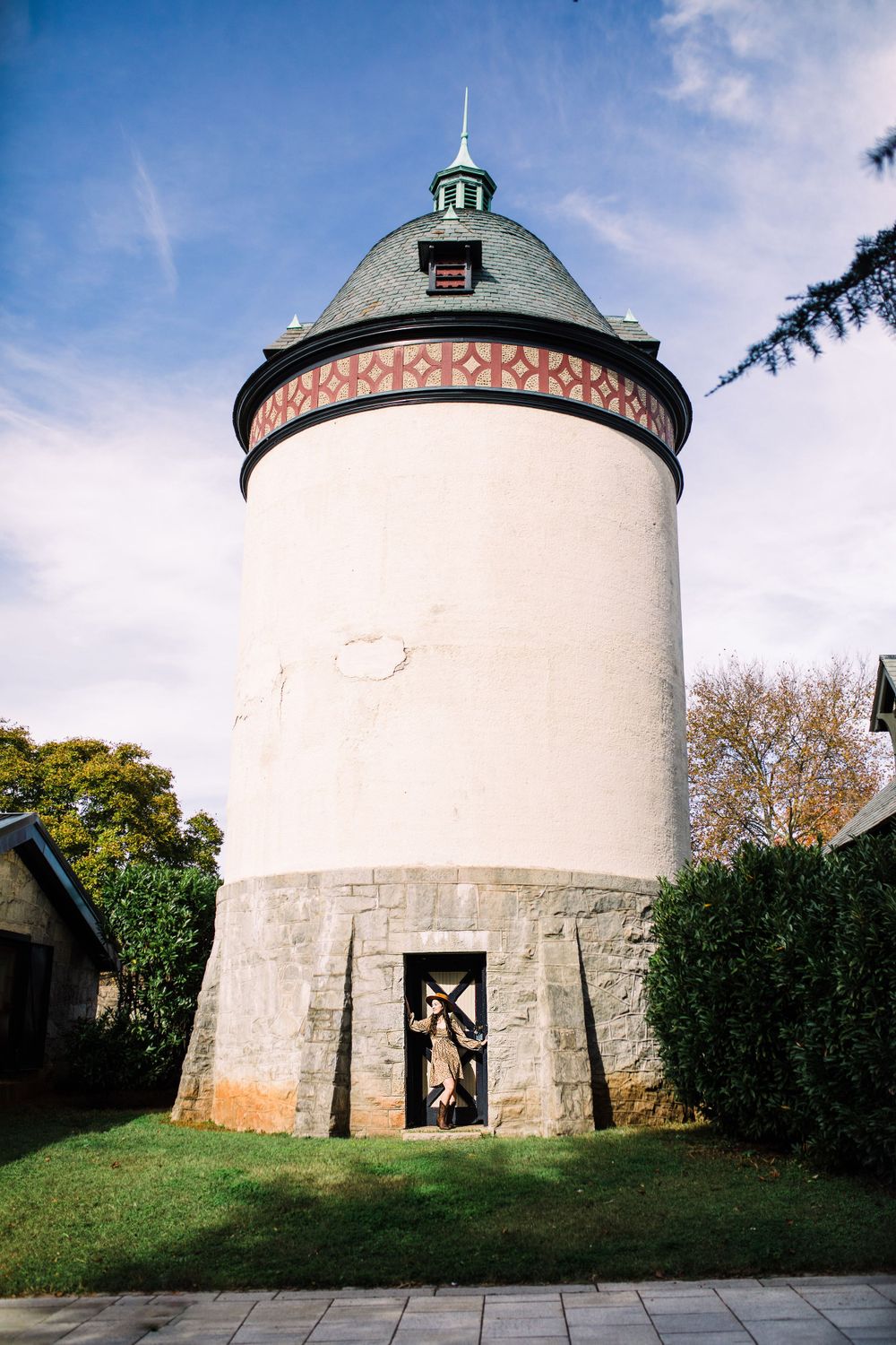 Historic round white tower with decorative red band and copper spire surrounded by green hedges.