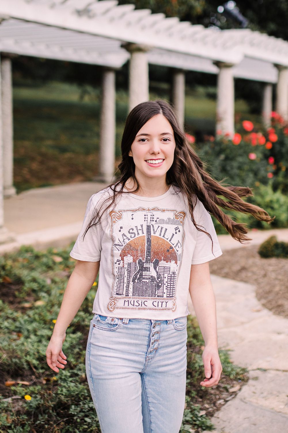 Young woman in Nashville Music City graphic tee walking through rose garden with hair blowing.