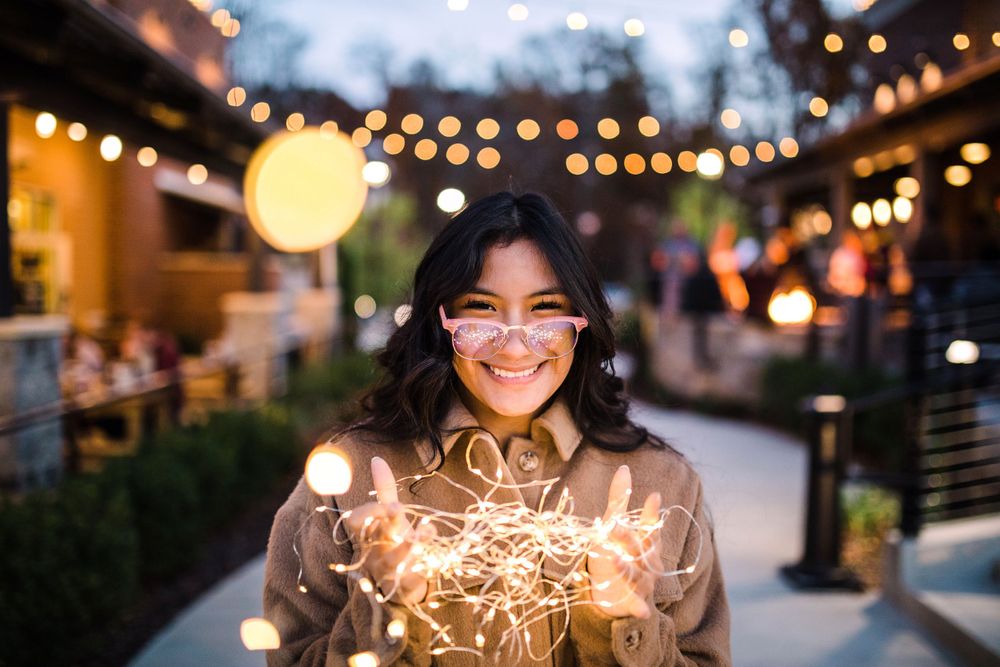 Happy young woman holding tangled fairy lights outdoors under string lights at dusk.