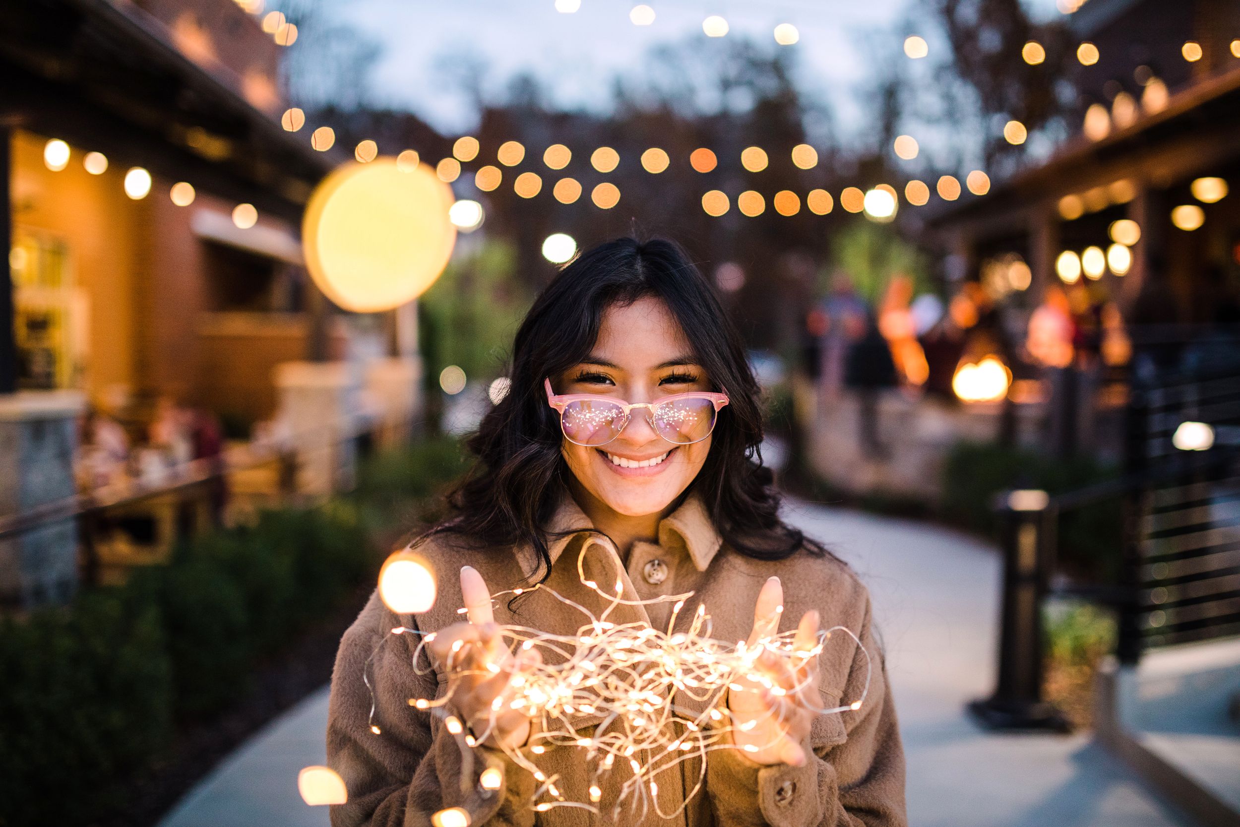 Happy young woman holding tangled fairy lights outdoors under string lights at dusk.