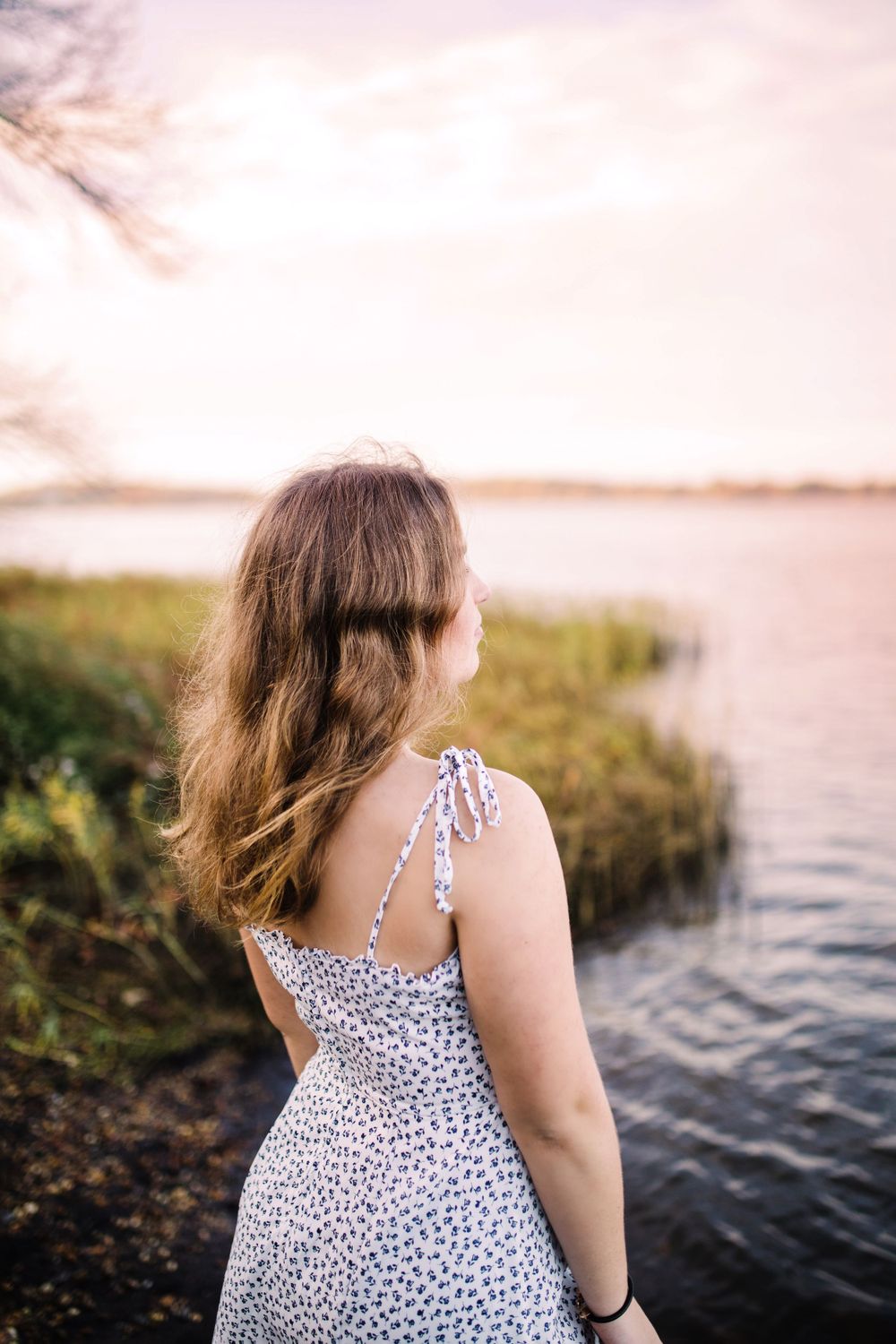 Young woman in white floral sundress viewed from behind gazing at peaceful lake at sunset.