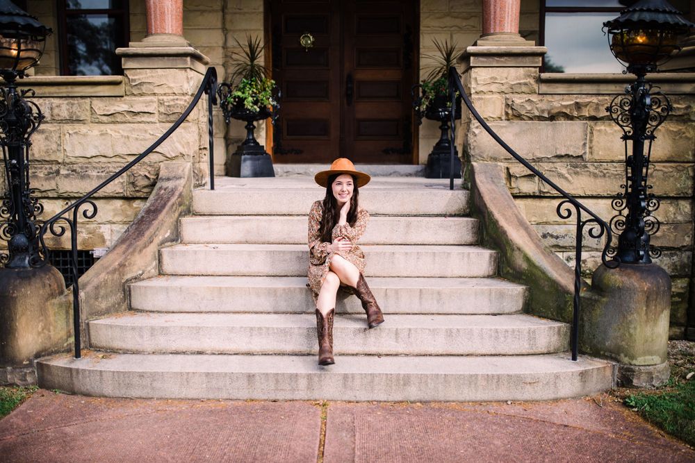 Young woman in brown hat and boots sitting on stone steps of historic building entrance.
