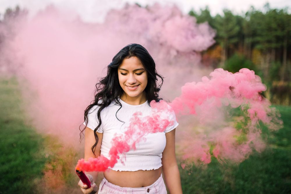 Smiling teen girl holding colorful pink smoke bomb in grassy field.