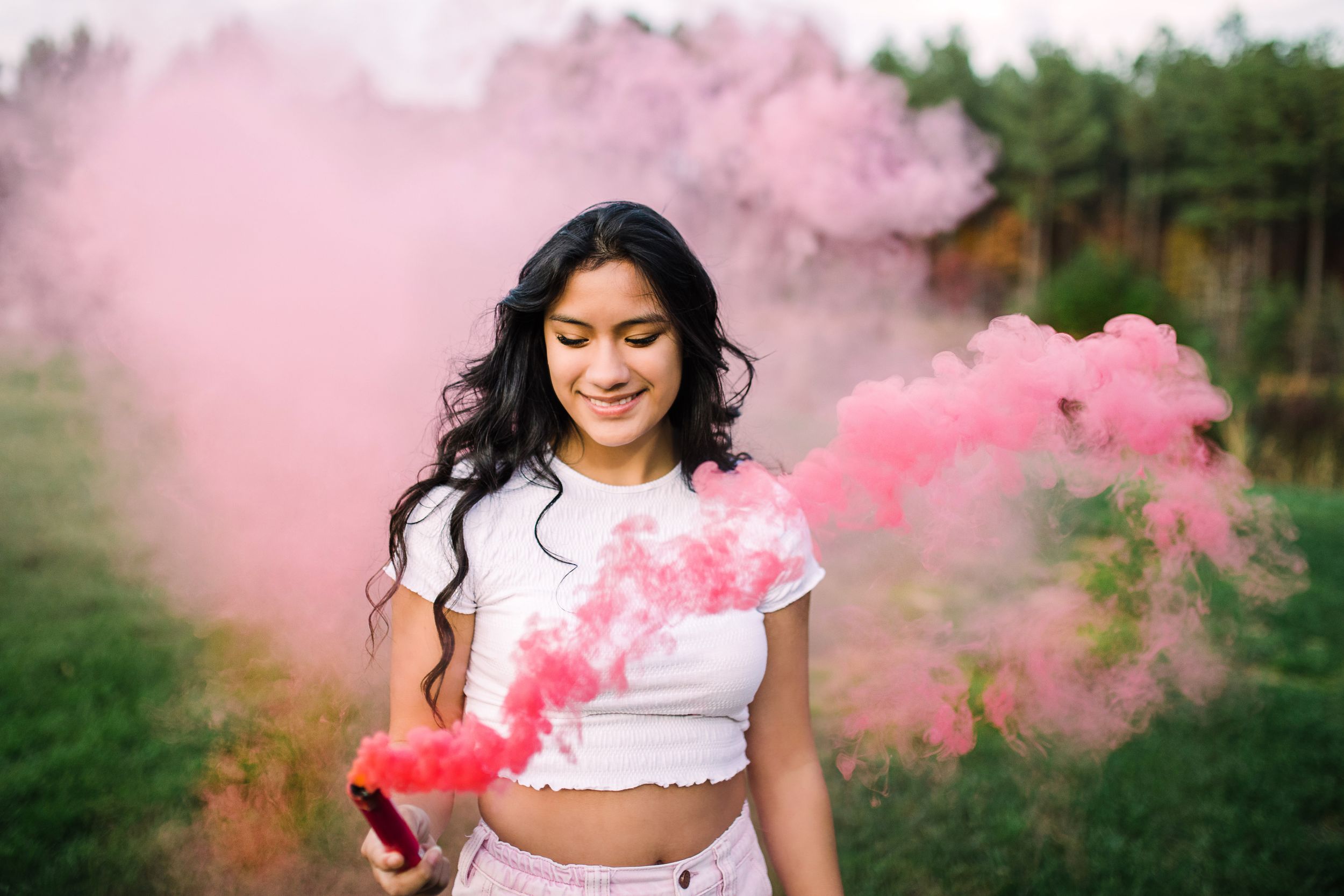 Smiling teen girl holding colorful pink smoke bomb in grassy field.