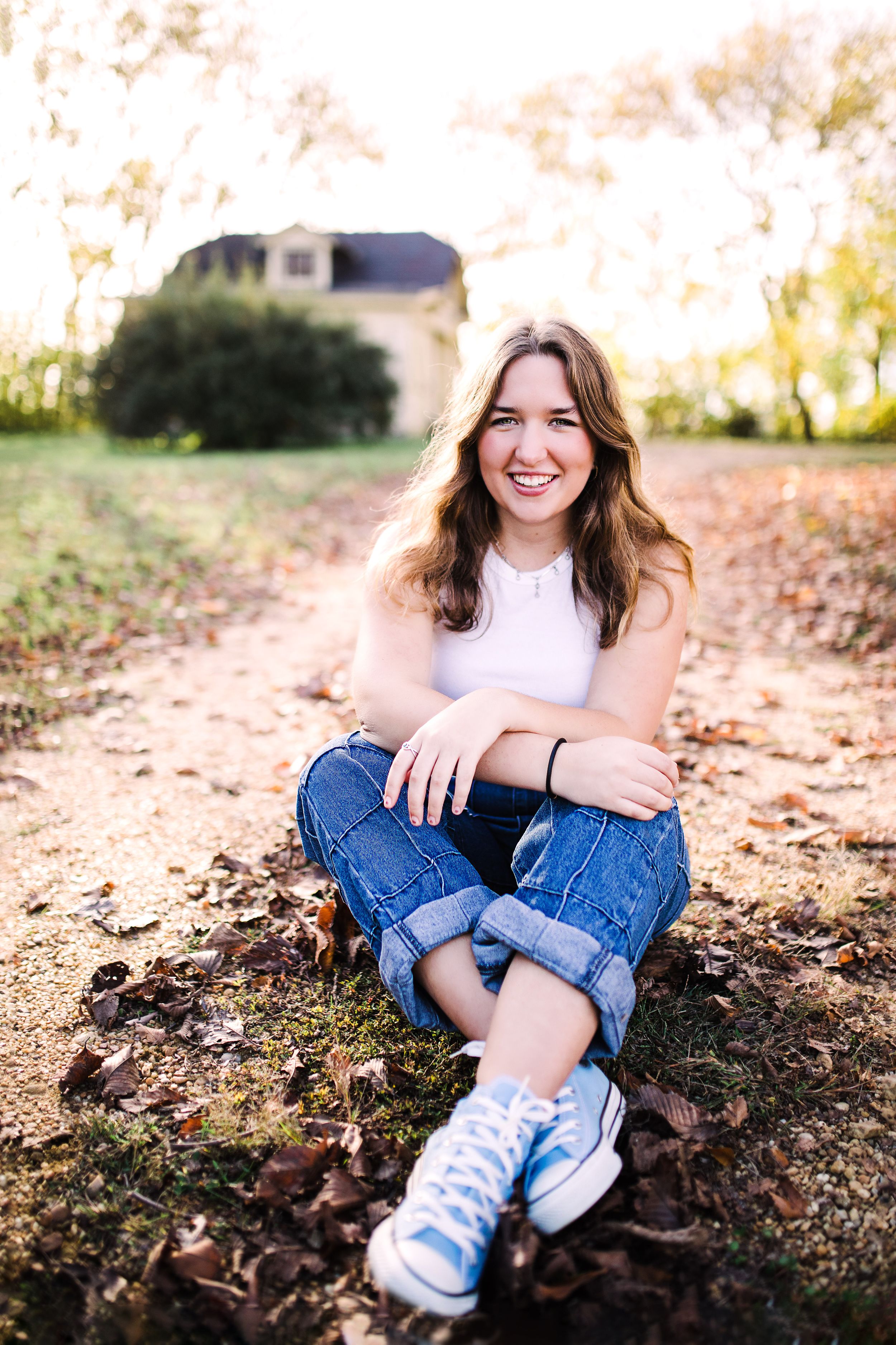 Happy young woman in white tank top and cuffed jeans sitting cross-legged on a leaf-covered ground.