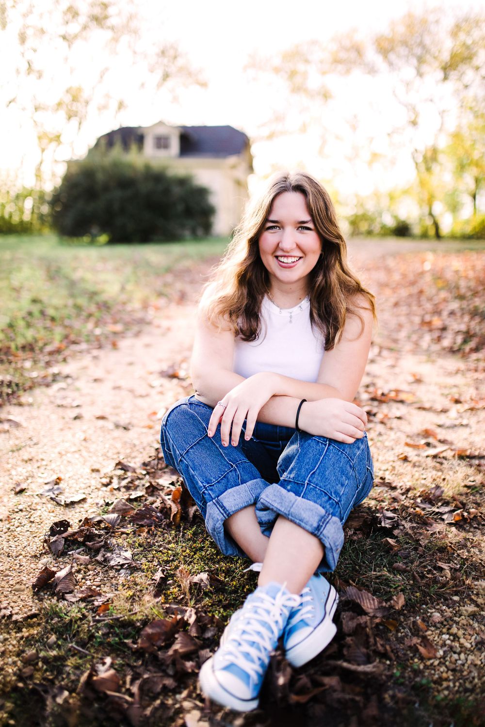 Happy young woman in white tank top and cuffed jeans sitting cross-legged on a leaf-covered ground.