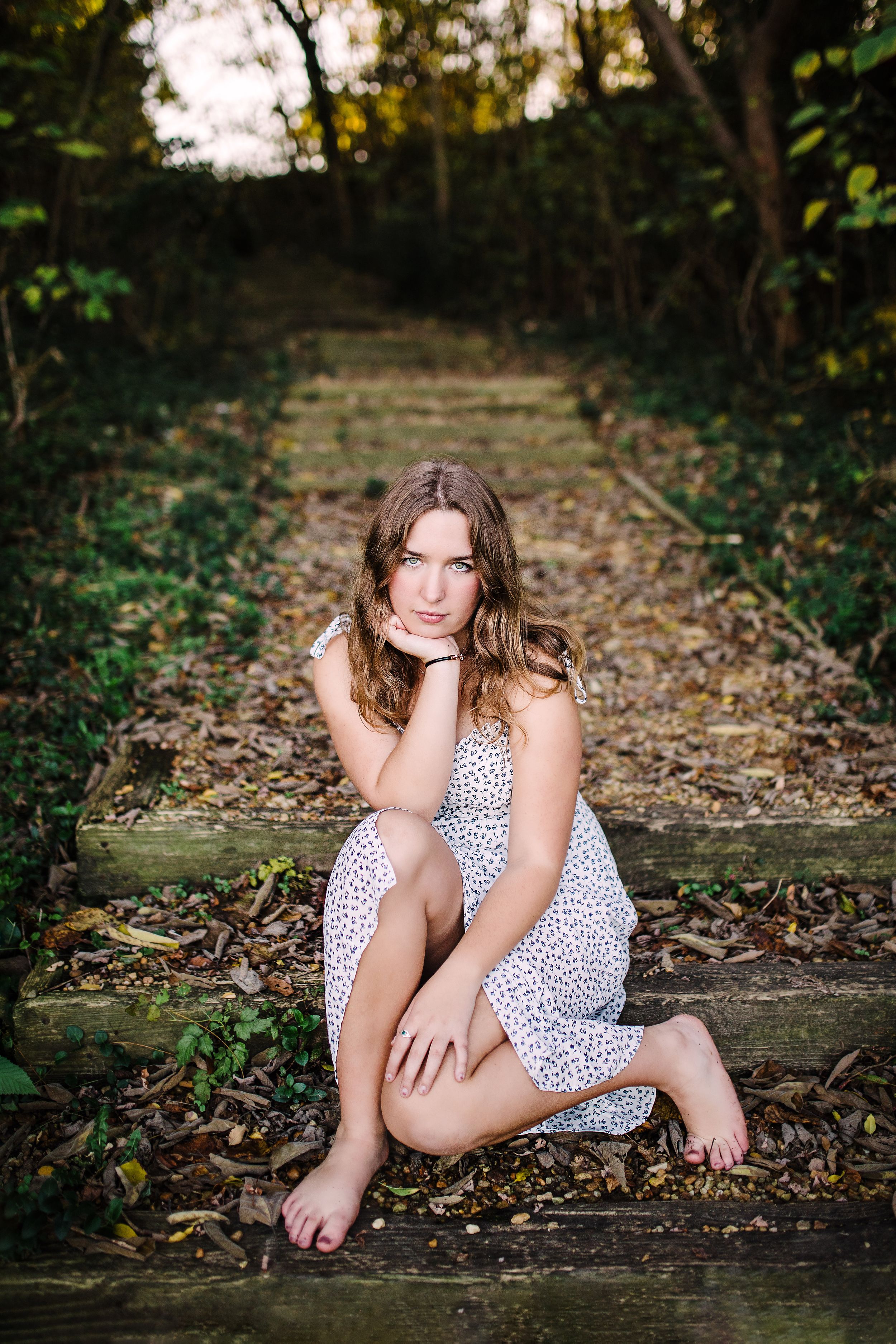 Young woman in white floral dress sitting barefoot on wooden outdoor steps surrounded by lush greenery.