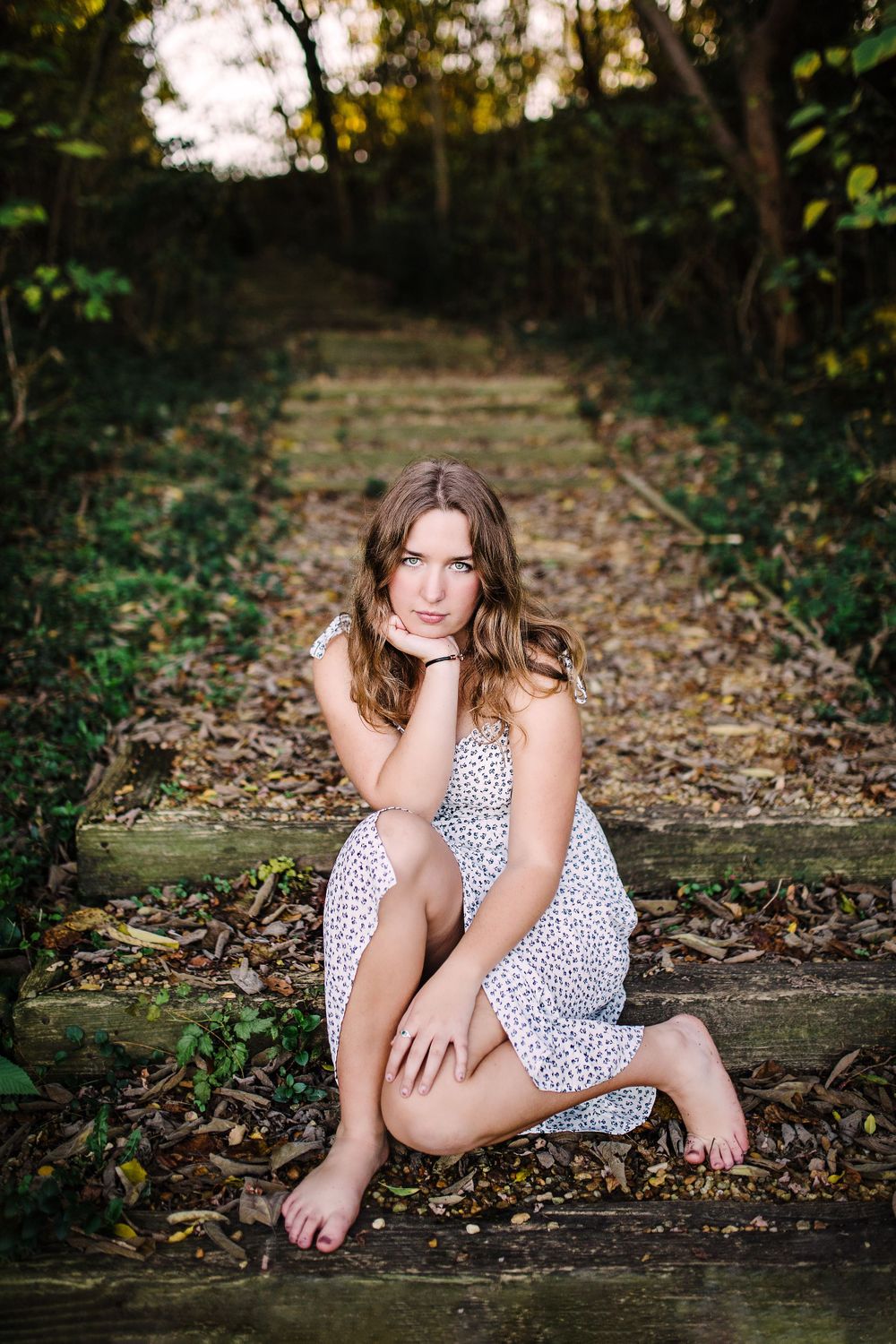 Young woman in white floral dress sitting barefoot on wooden outdoor steps surrounded by lush greenery.