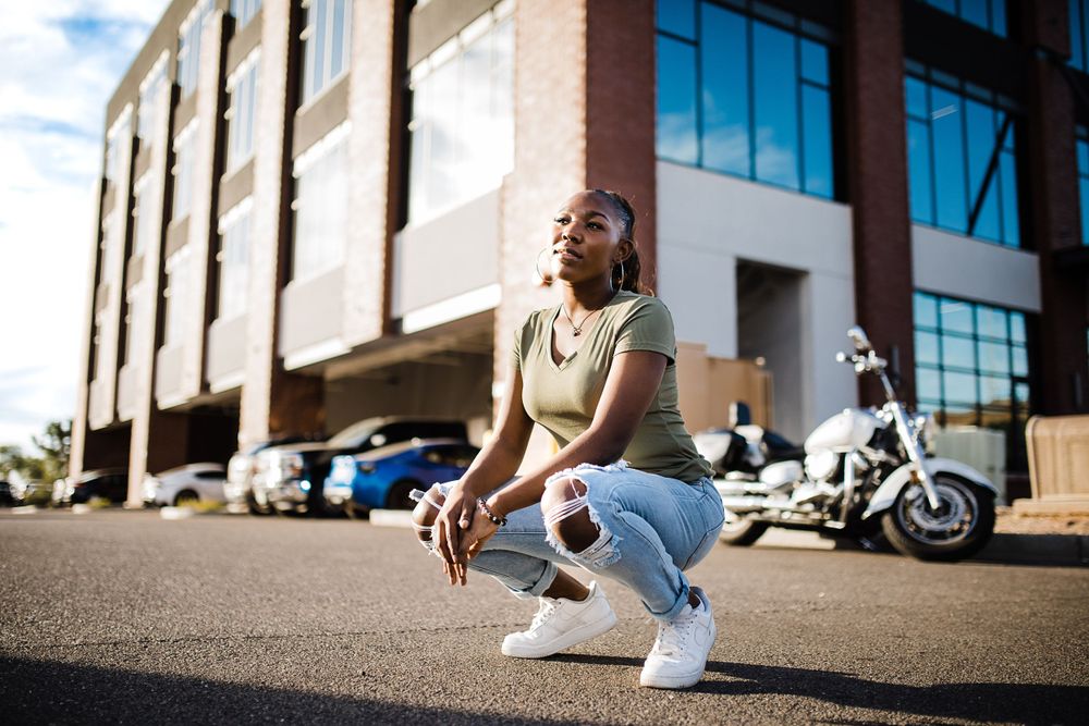 Young Black woman in olive top and jeans crouches in parking lot with motorcycle and building behind her.