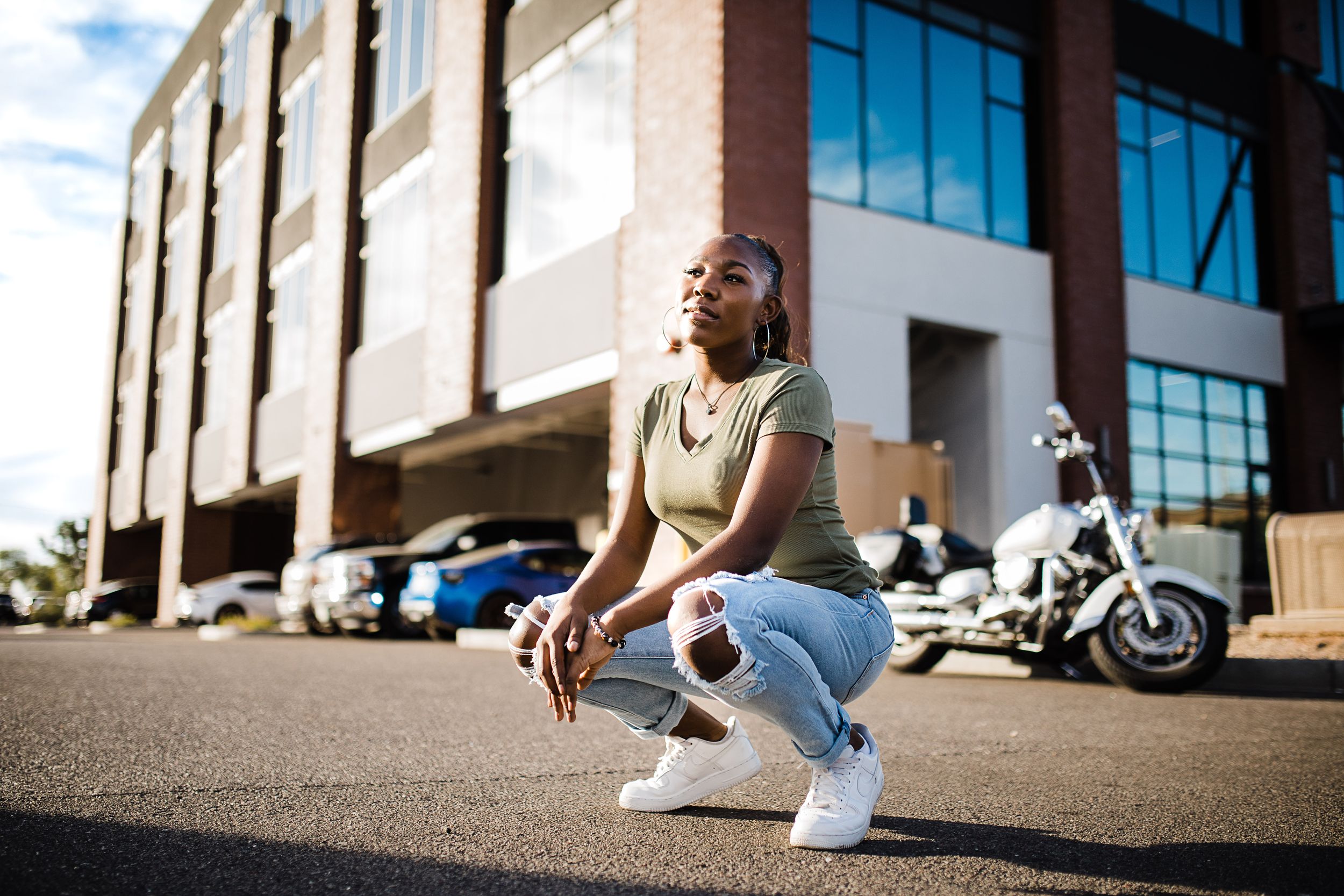 Young Black woman in olive top and jeans crouches in parking lot with motorcycle and building behind her.