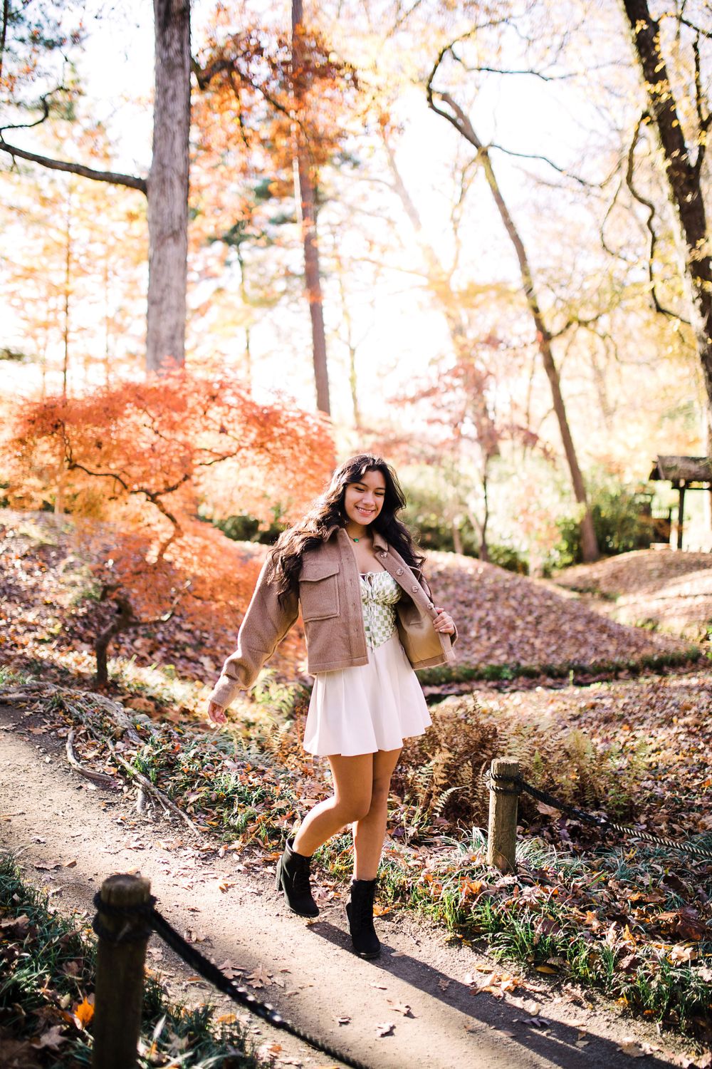 Teen girl in white dress and tan jacket walking on path through colorful autumn forest.