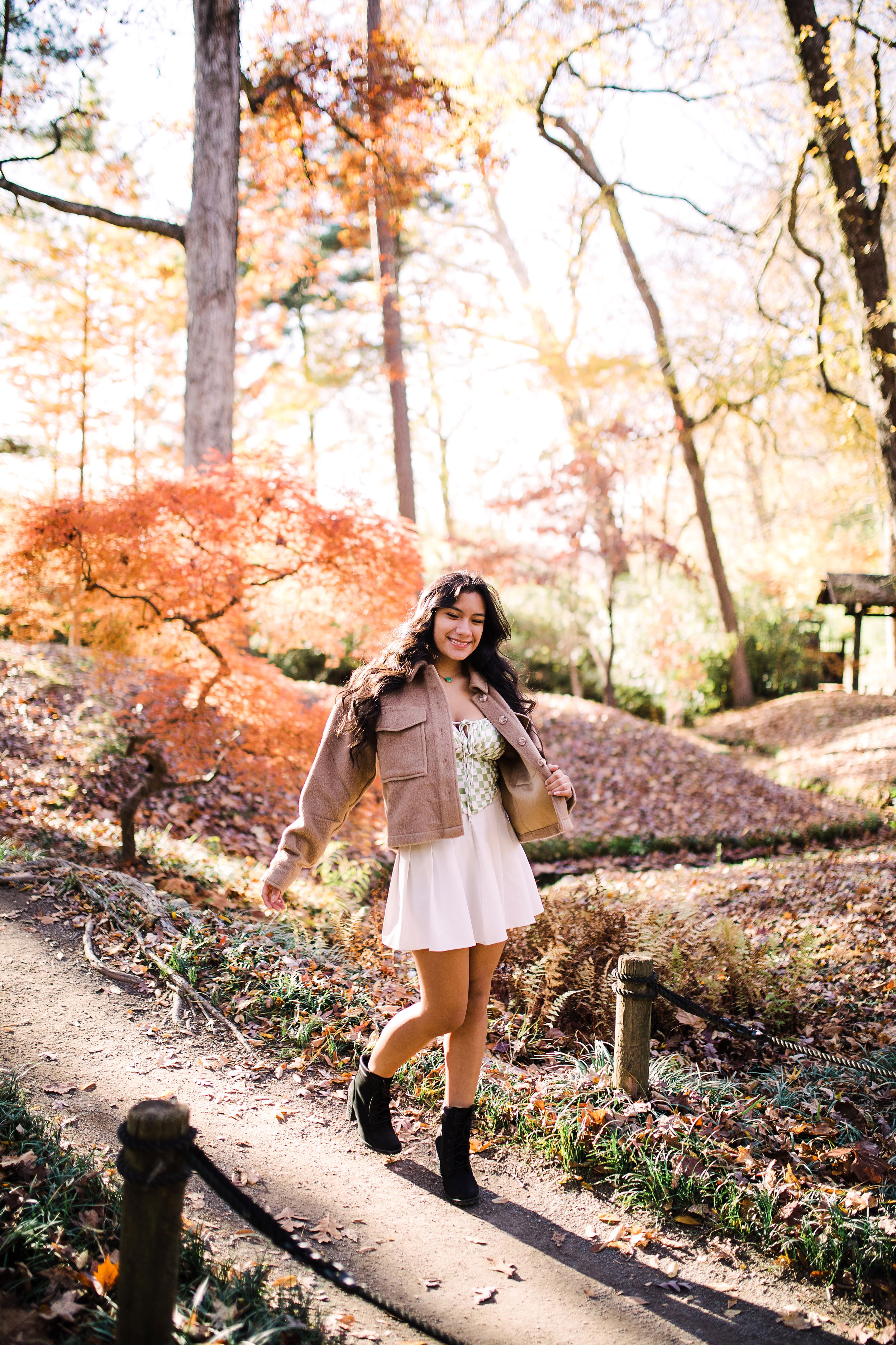 Teen girl in white dress and tan jacket walking on path through colorful autumn forest.