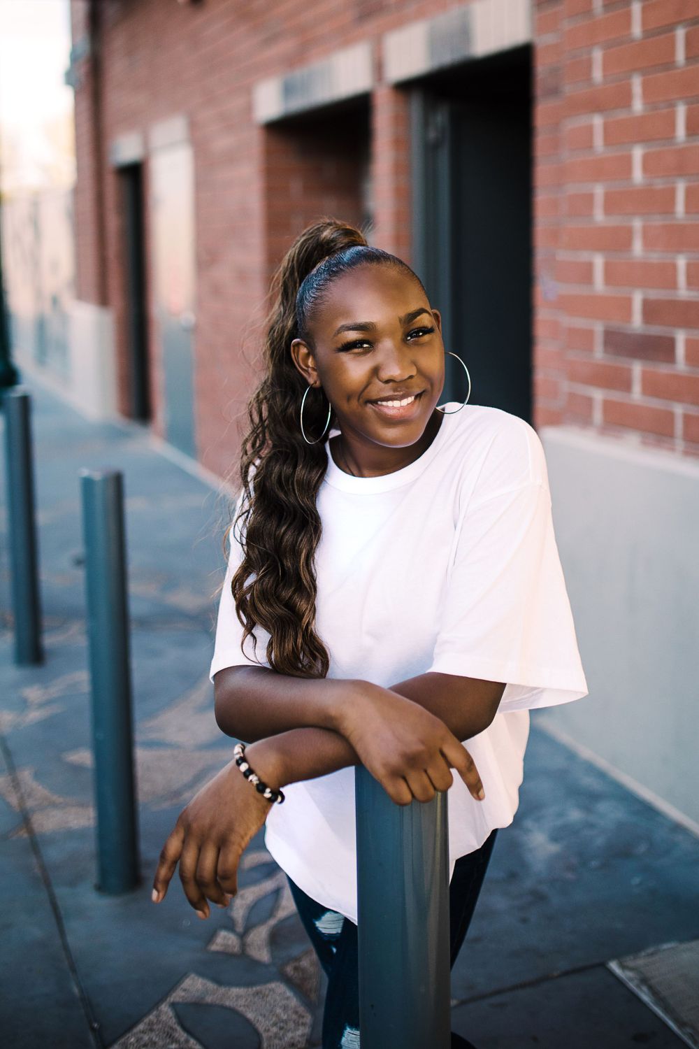 Smiling young Black woman with long ponytail leans on pole in urban alley wearing white oversized tee.