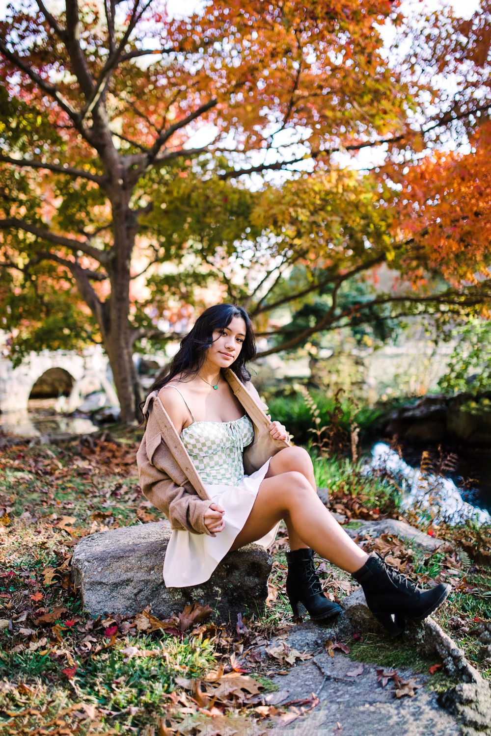 Young woman in white dress and tan coat sits on a rock surrounded by colorful fall leaves.