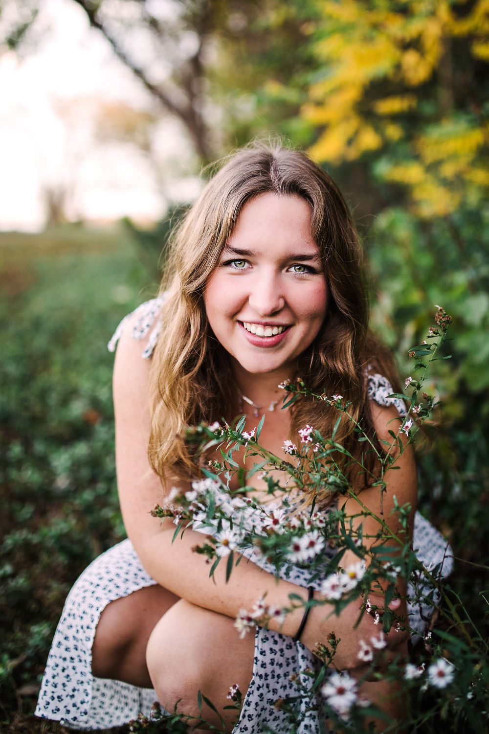 Smiling young woman with blue eyes in white dress holding wild pink flowers in green garden.