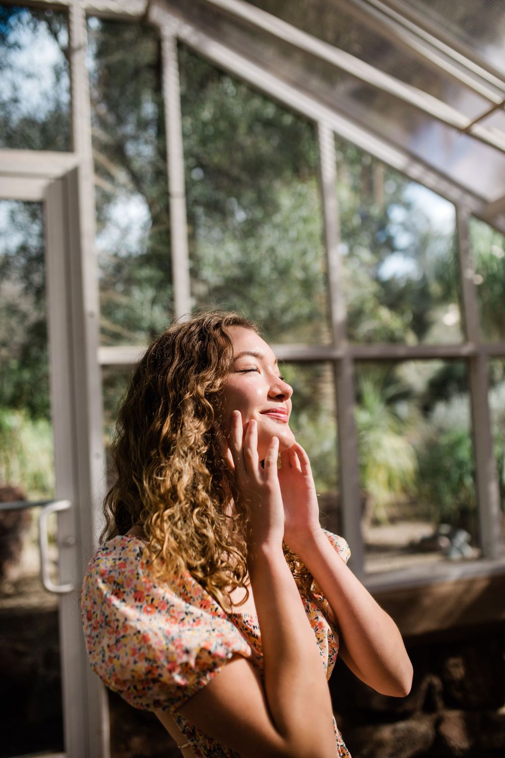 Curly-haired young woman in floral dress enjoying sunlight with eyes closed near greenhouse windows.