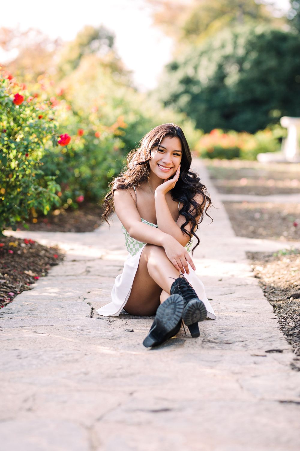Smiling young woman with long wavy hair sits on stone path surrounded by blooming roses in a garden.