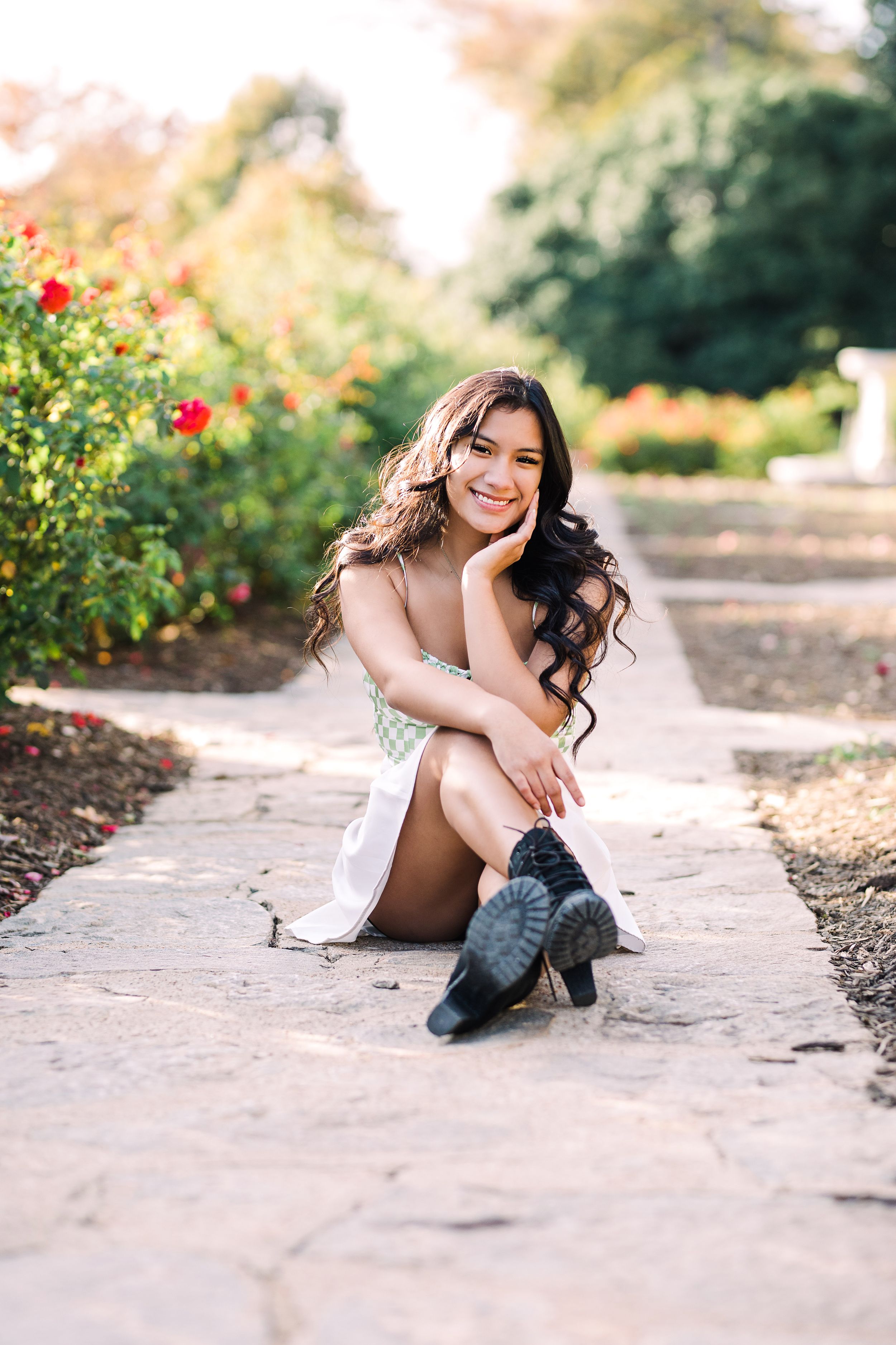Smiling young woman with long wavy hair sits on stone path surrounded by blooming roses in a garden.