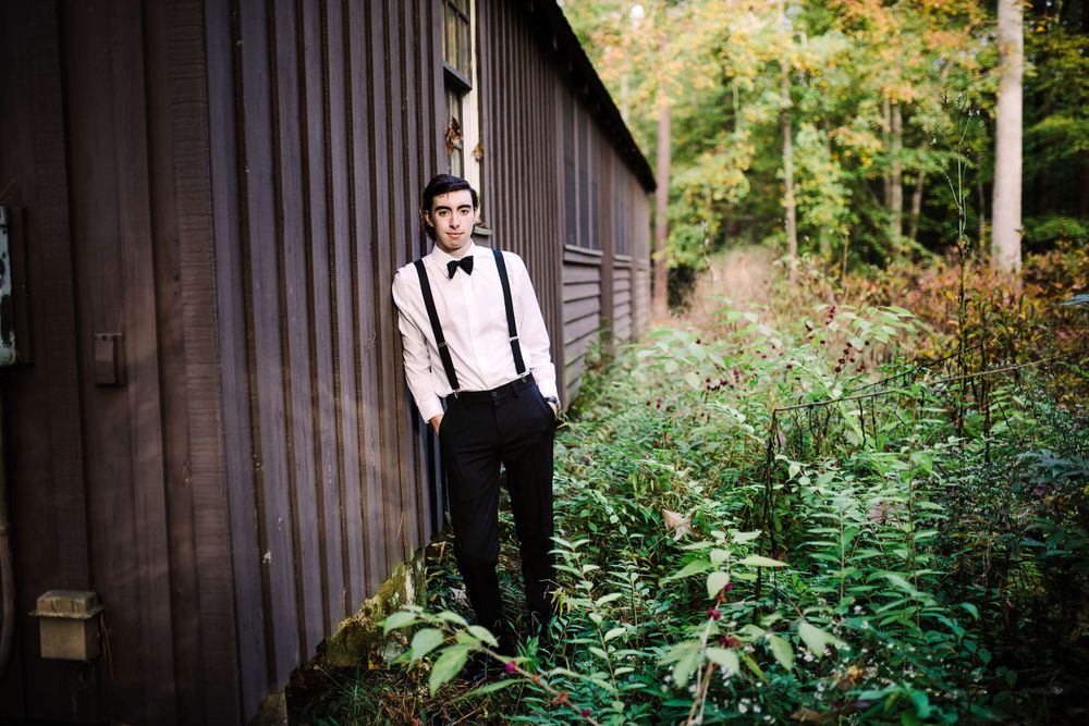 Stylish young man in white shirt, suspenders and bow tie standing by dark wooden building in forest.