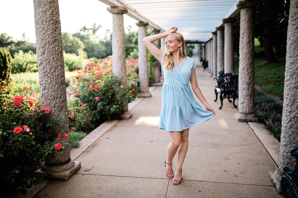 Blonde teen posing in light blue dress among stone columns with red roses in background.