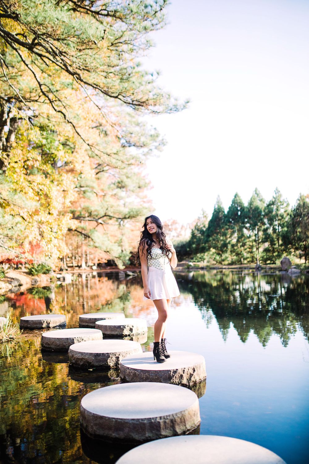 Young woman in white dress and black boots stands on stepping stones over a serene autumn lake.