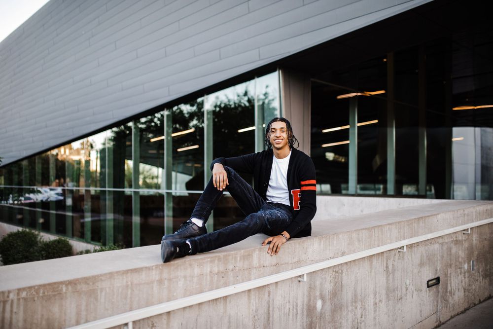 Young man in black varsity cardigan sits casually on concrete ledge outside modern glass building.