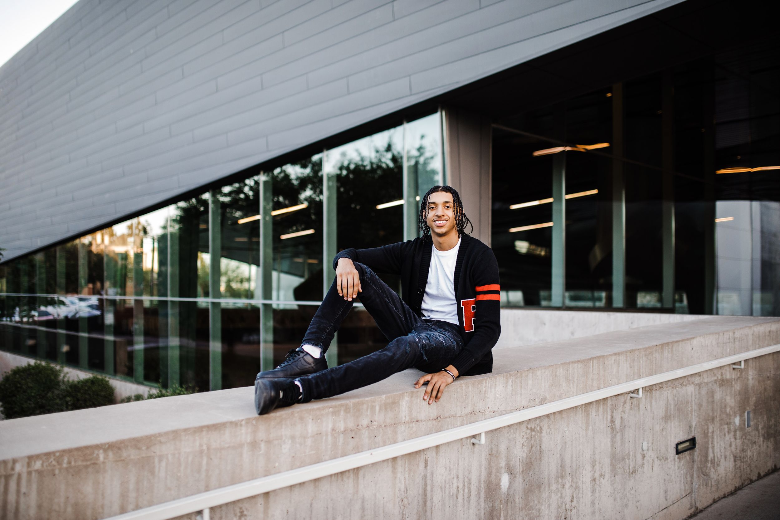 Young man in black varsity cardigan sits casually on concrete ledge outside modern glass building.