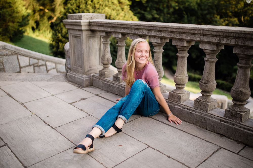 Smiling blonde teen in pink top and jeans sitting on stone terrace with balustrade.